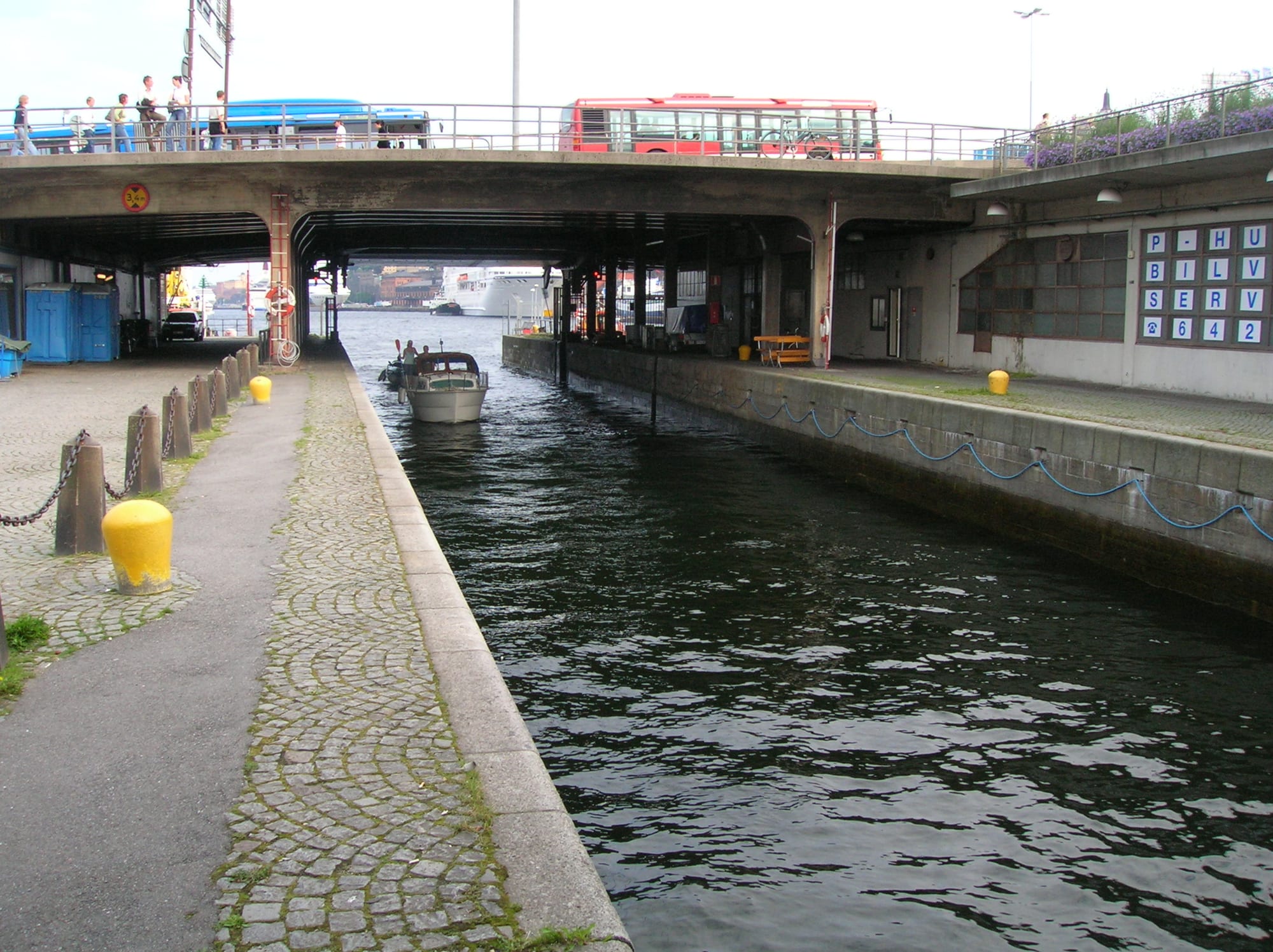 A small motor boat waiting in a narrow stone lock channel beneath a road bridge carrying a red bus