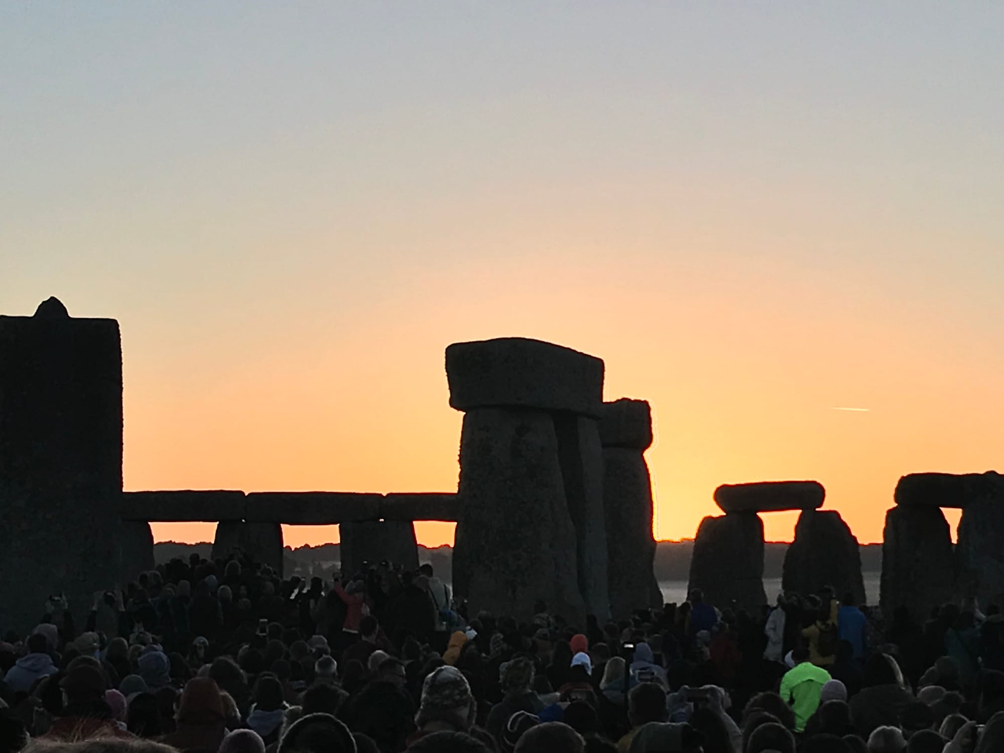 The silhouettes of massive standing stones and lintels against an orange dawn sky, with a dense crowd of people in the foreground