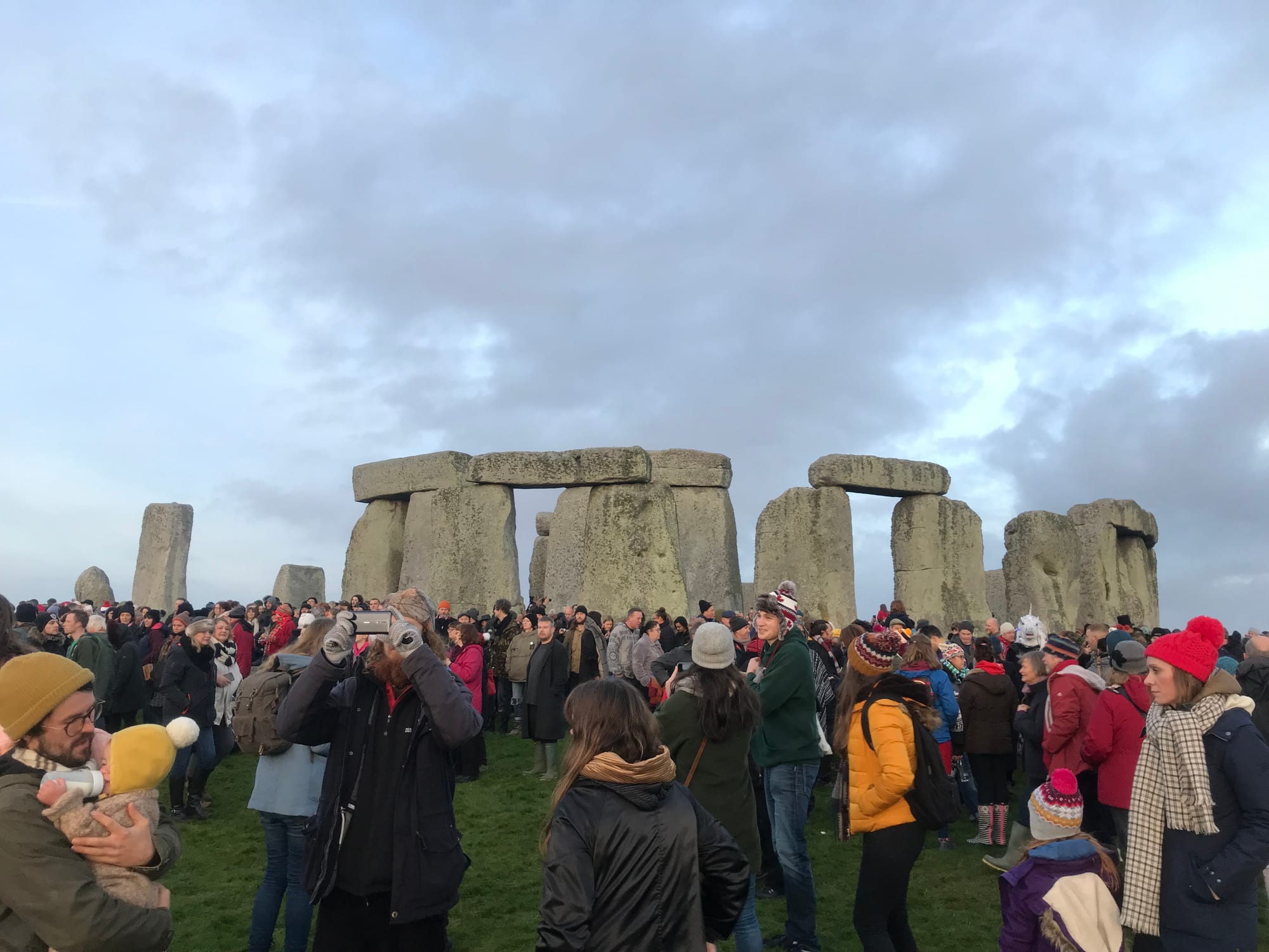 A crowd of people in winter coats, scarves and woolly hats stands among the great sarsen stones and lintels of Stonehenge under a grey sky