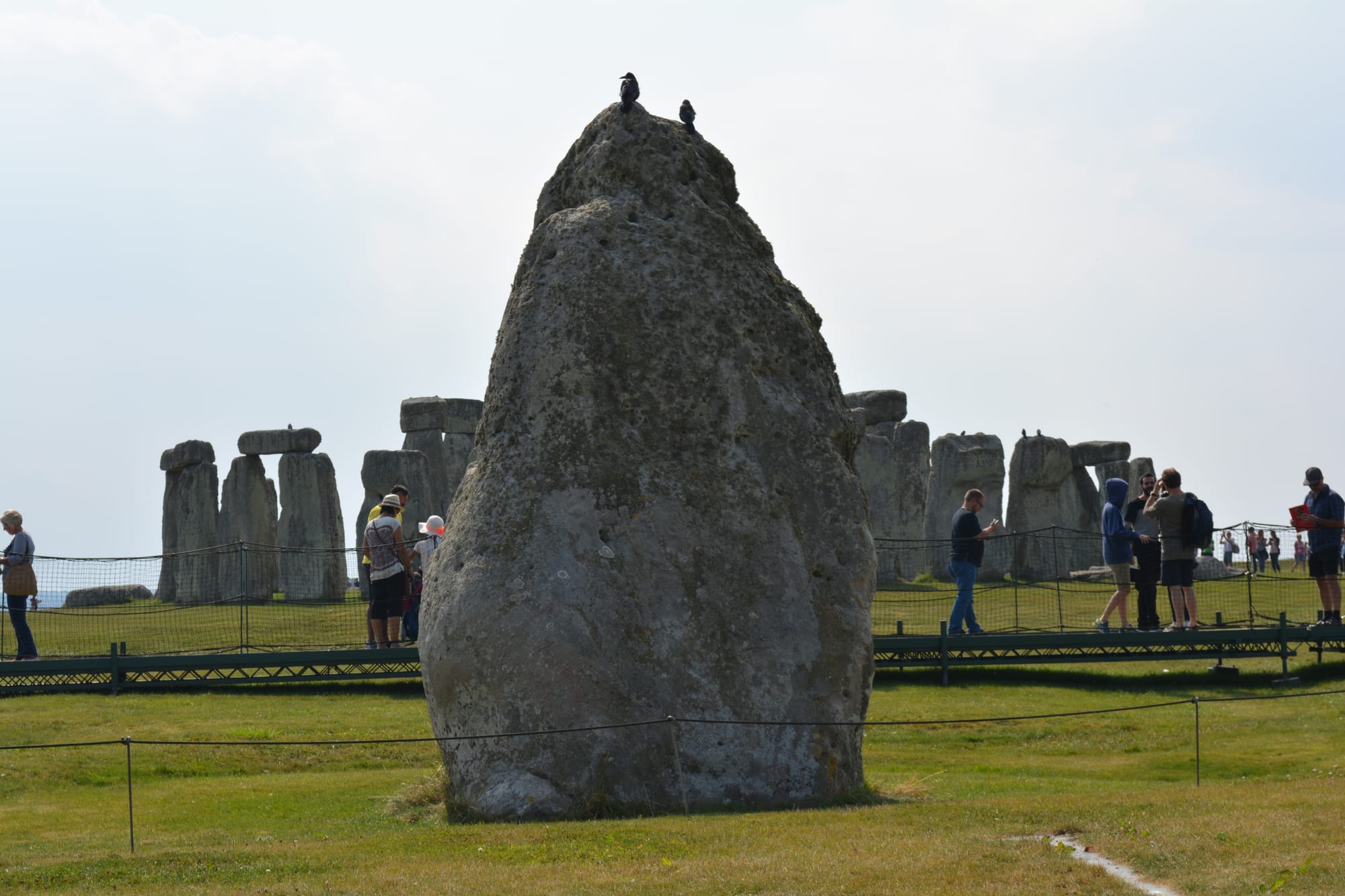 A huge, rough, leaning standing stone on a lawn, with the circle of sarsen stones and lintels behind it and visitors walking around