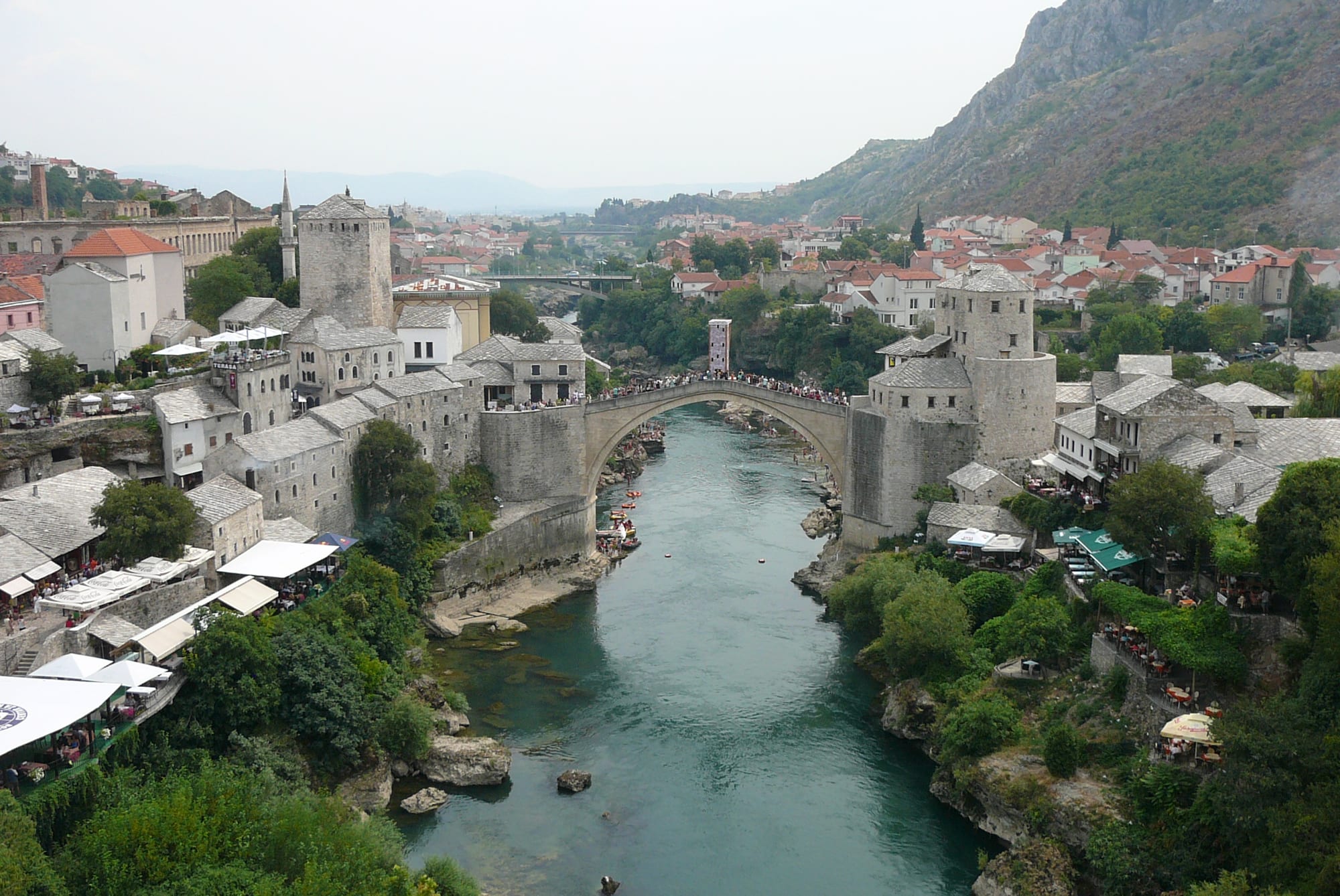 A single high stone arch between two stone towers spanning a green river, with old stone houses and a rocky mountainside around it