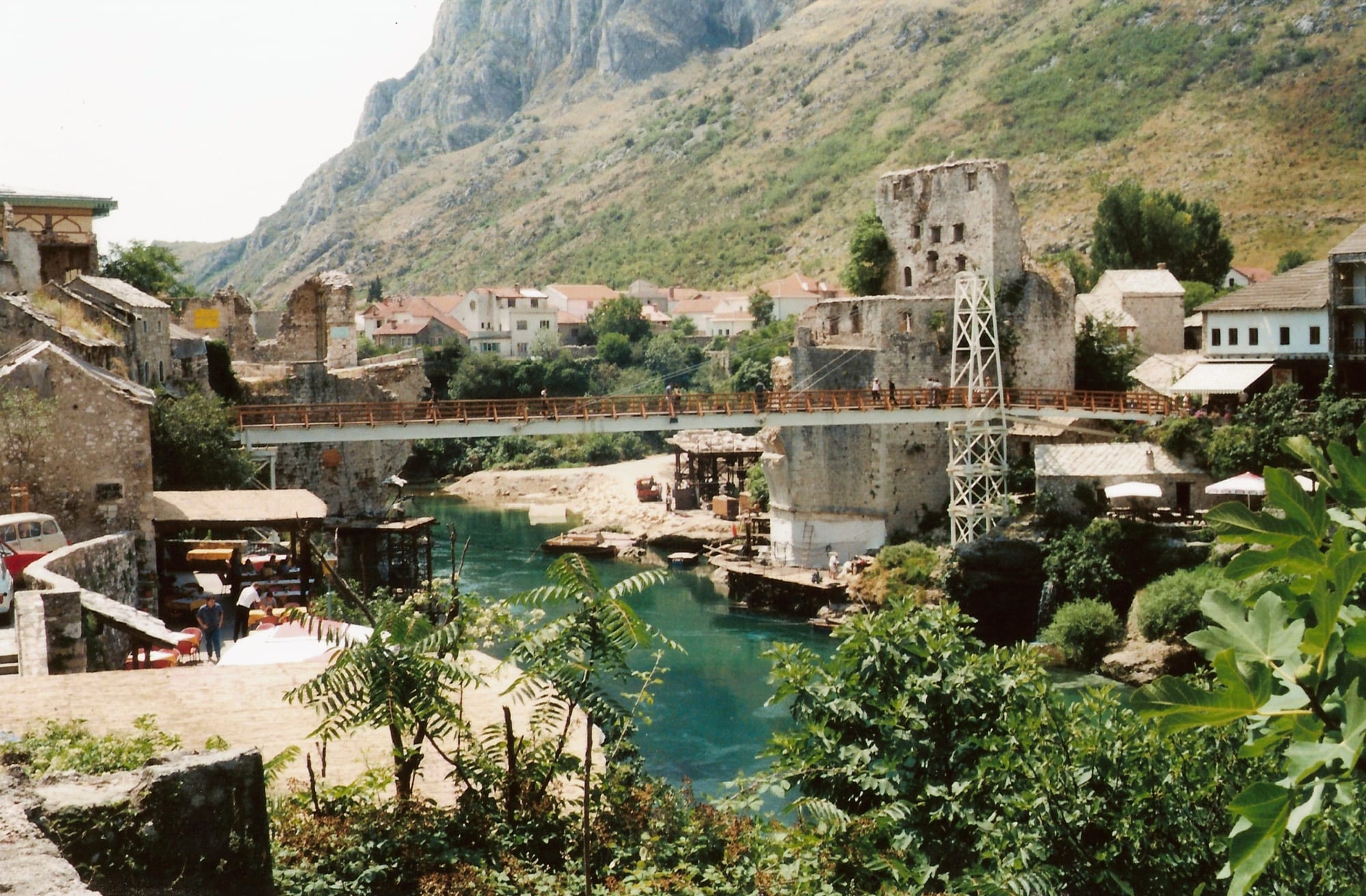 The ruined stumps of a stone bridge on either bank of a green river, with a temporary metal footbridge strung across between them