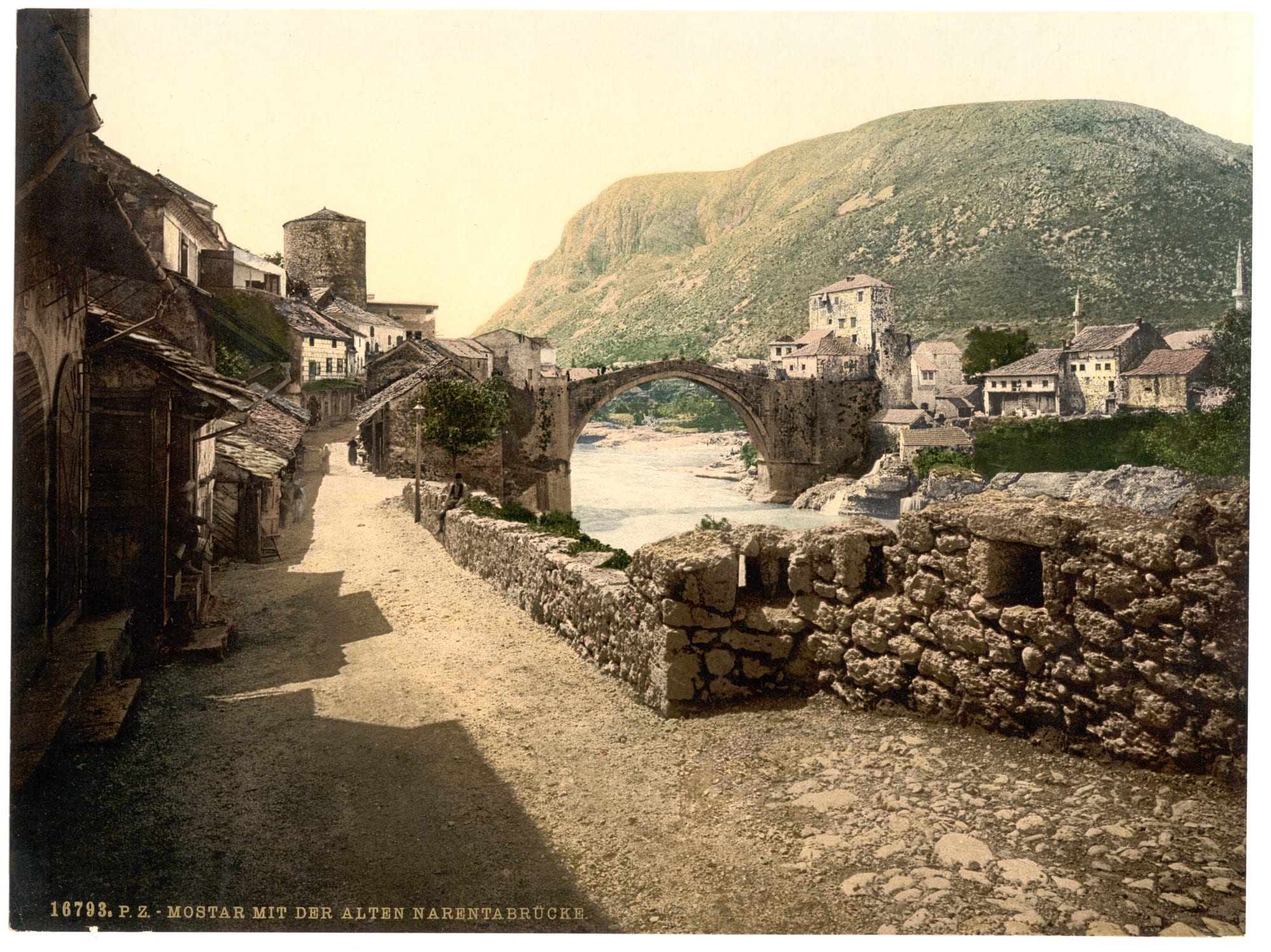 A hand-coloured old photograph of a stone arch bridge over a pale river, with a dusty lane, stone houses and a bare hillside