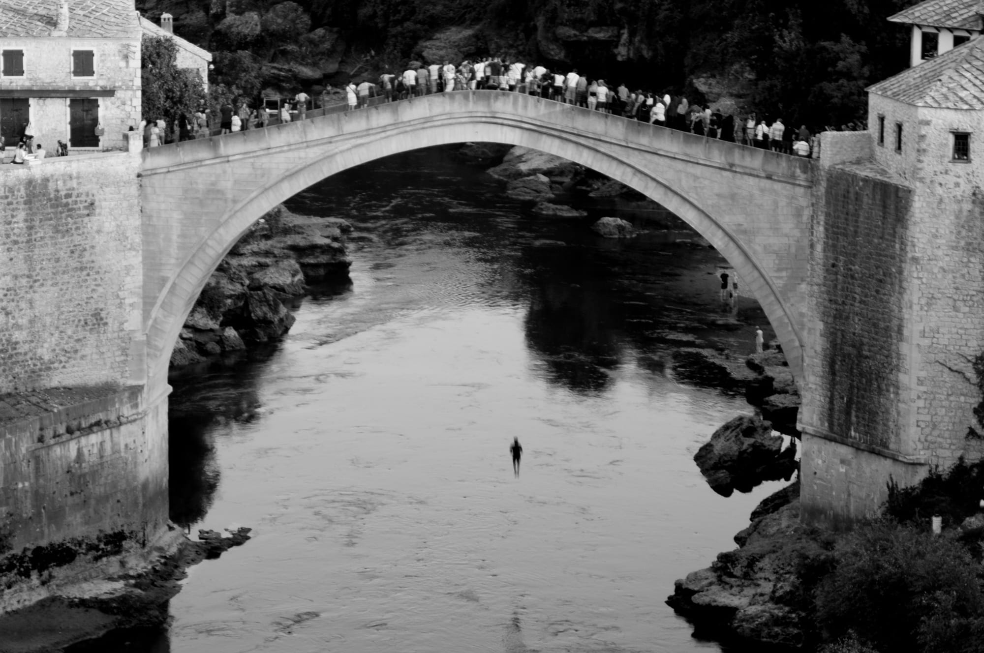 A black and white photograph of a diver in mid-air beneath a stone arch packed with spectators, above a dark river