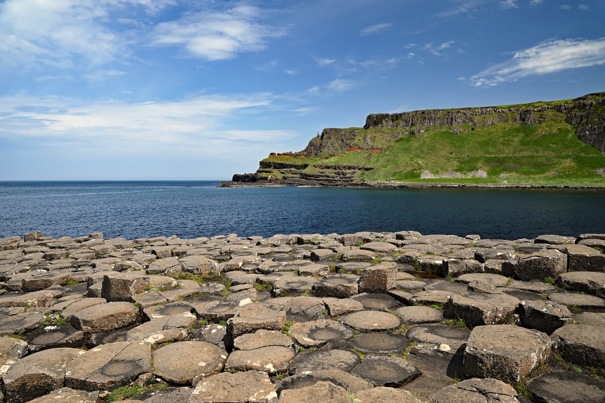Flat-topped, many-sided stone columns packed together like paving at the edge of a calm blue bay, with green headlands and dark cliffs beyond