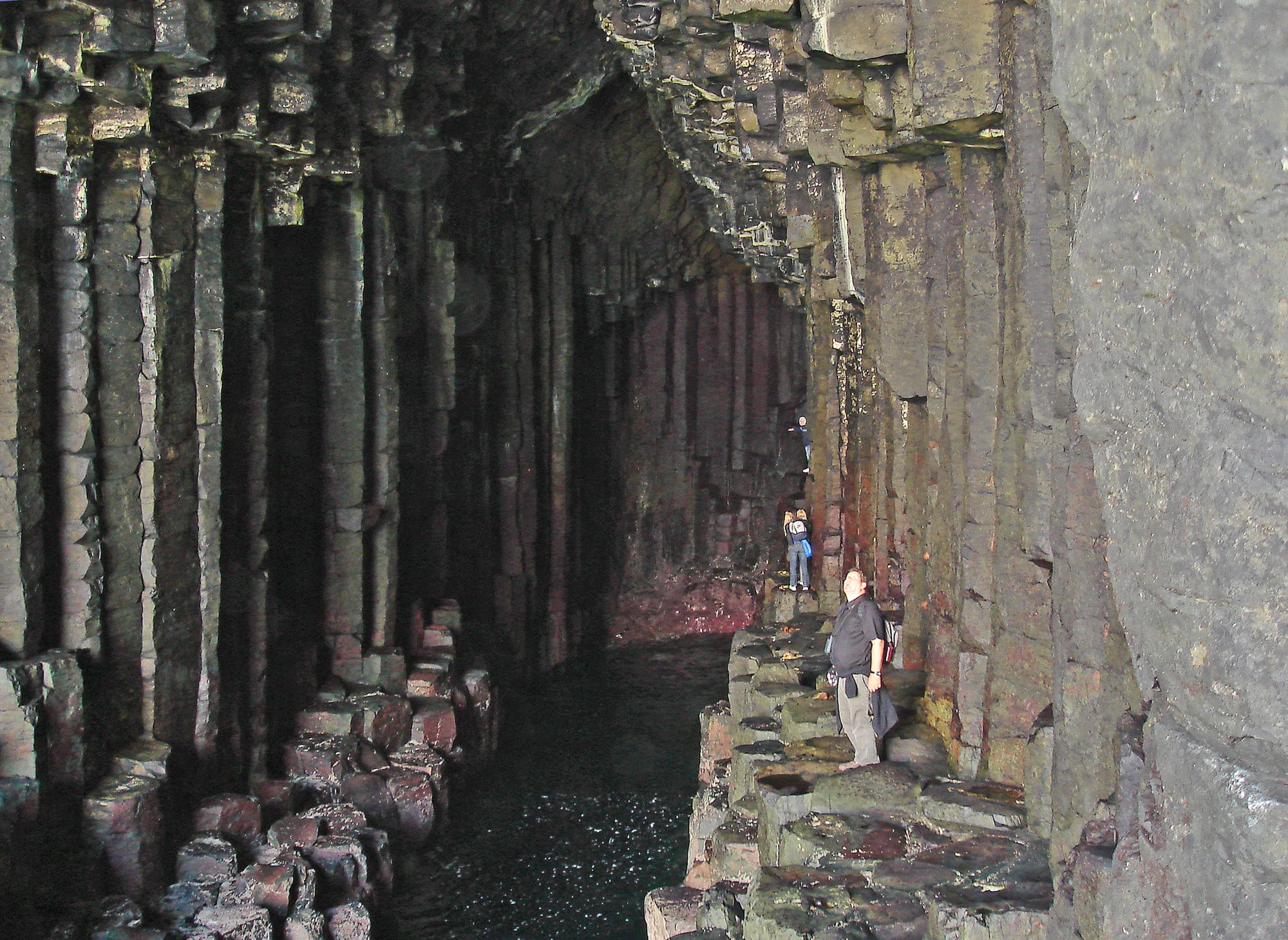 A tall sea cave whose walls are made of vertical stone columns, with two visitors standing on broken column tops beside dark water