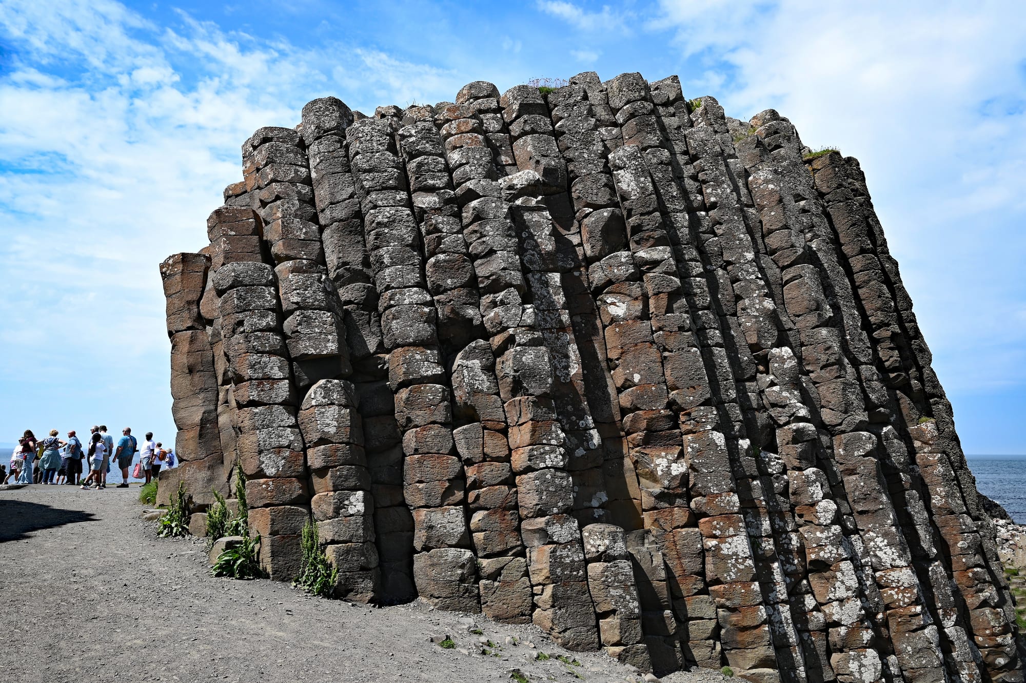 A tall block of stone made of many columns, each split horizontally into short stacked segments, with visitors walking past