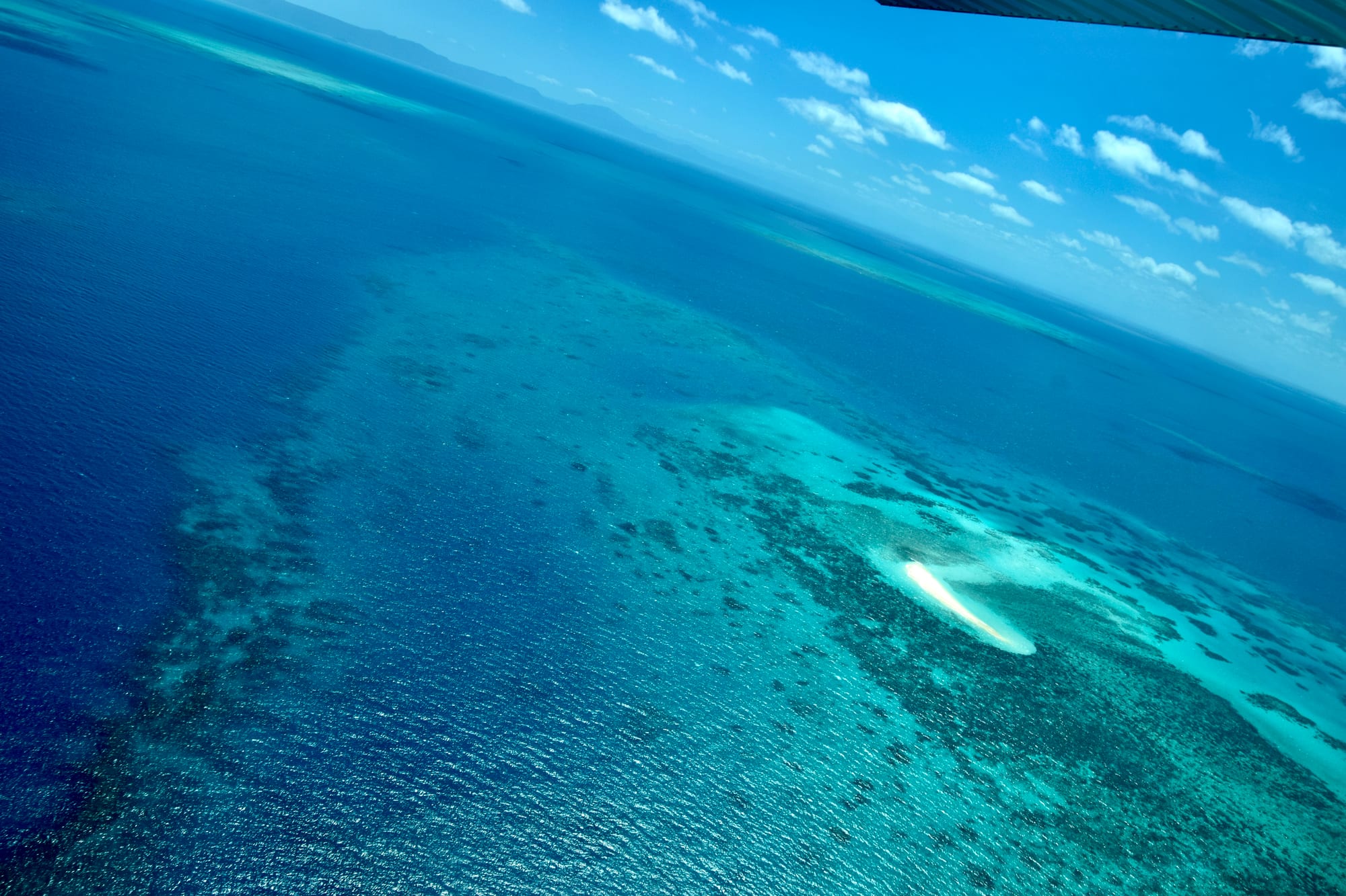 An aerial view of turquoise shallows, dark coral patches and a small white sand cay in a deep blue sea, with more reefs stretching to the horizon