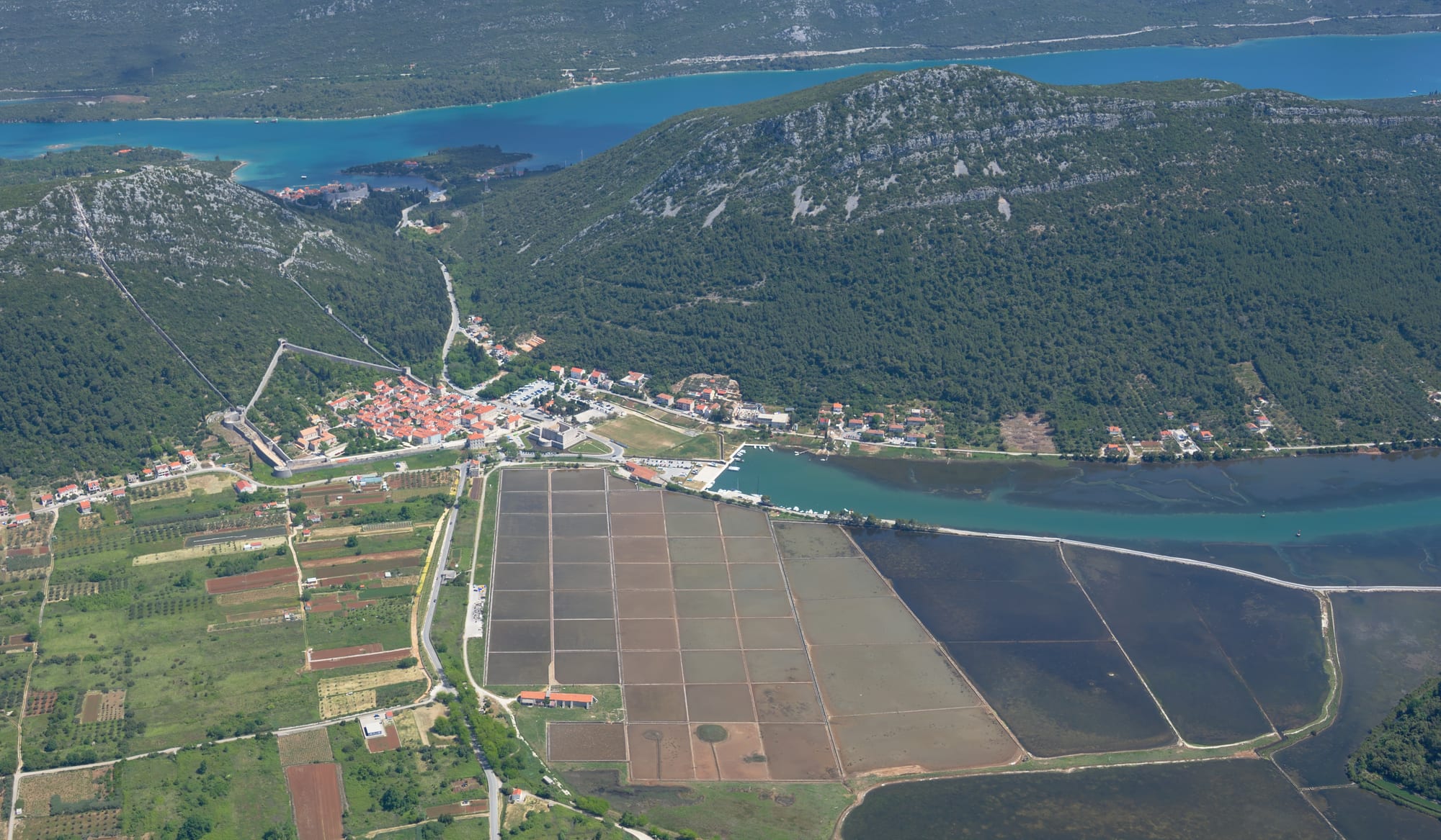 An aerial view of a grid of flat rectangular salt ponds beside a small walled town, green hills and a blue inlet of the sea