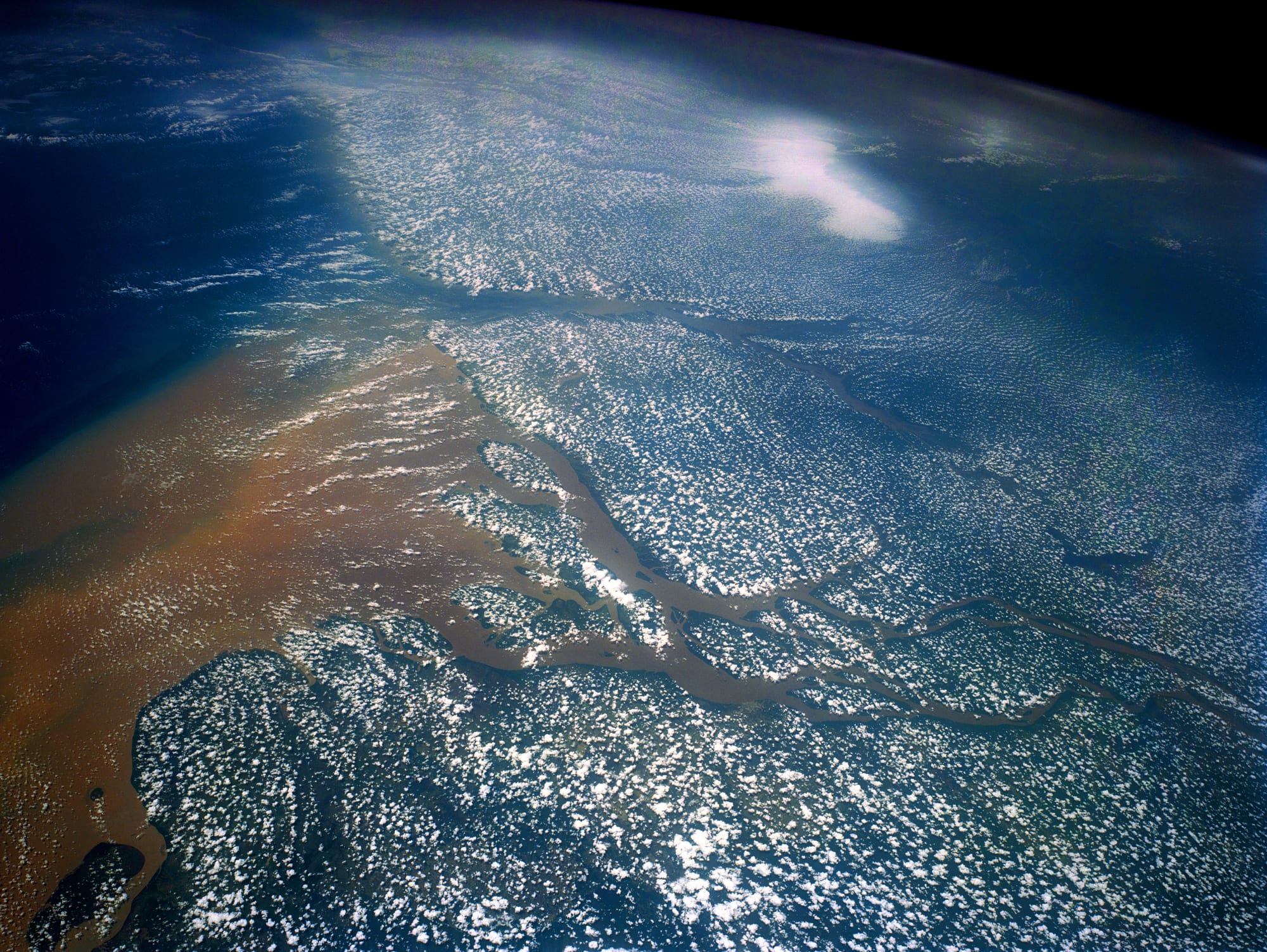 A view from orbit of a huge river splitting into many channels as it meets the sea, with a broad brown plume of sediment spreading into the blue ocean