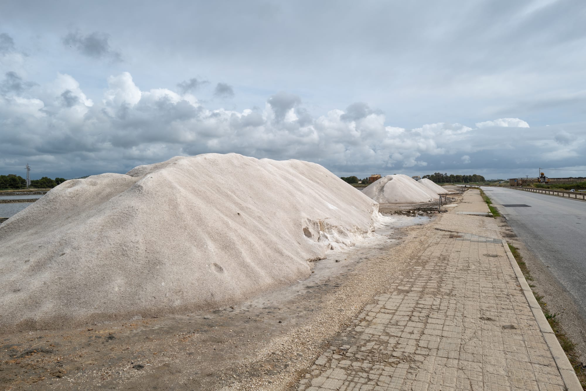 Large white heaps of salt beside a paved path and a road, with flat salt pans and a cloudy sky behind