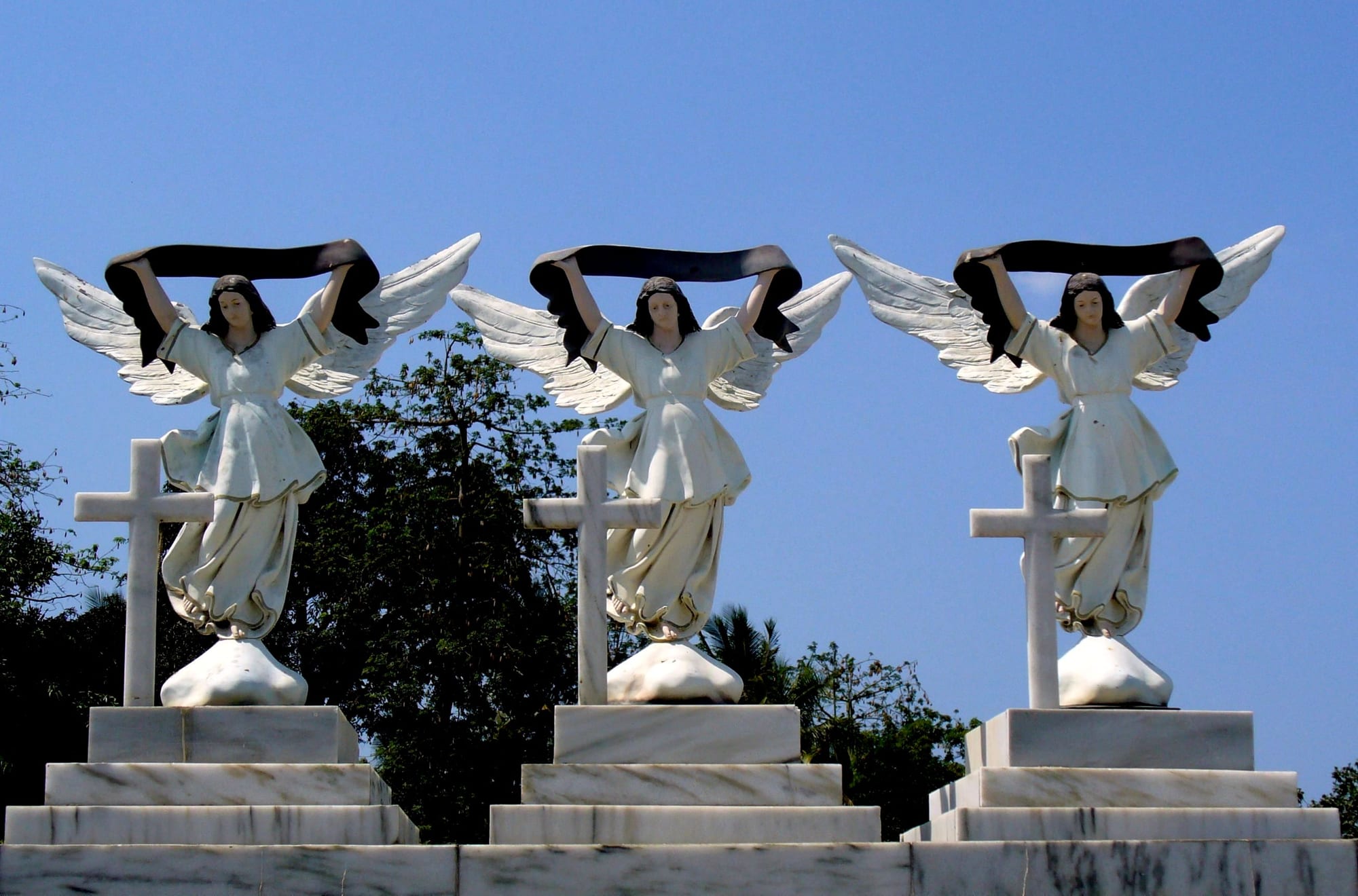 Three white marble angel statues holding black sashes above white crosses, against a blue sky