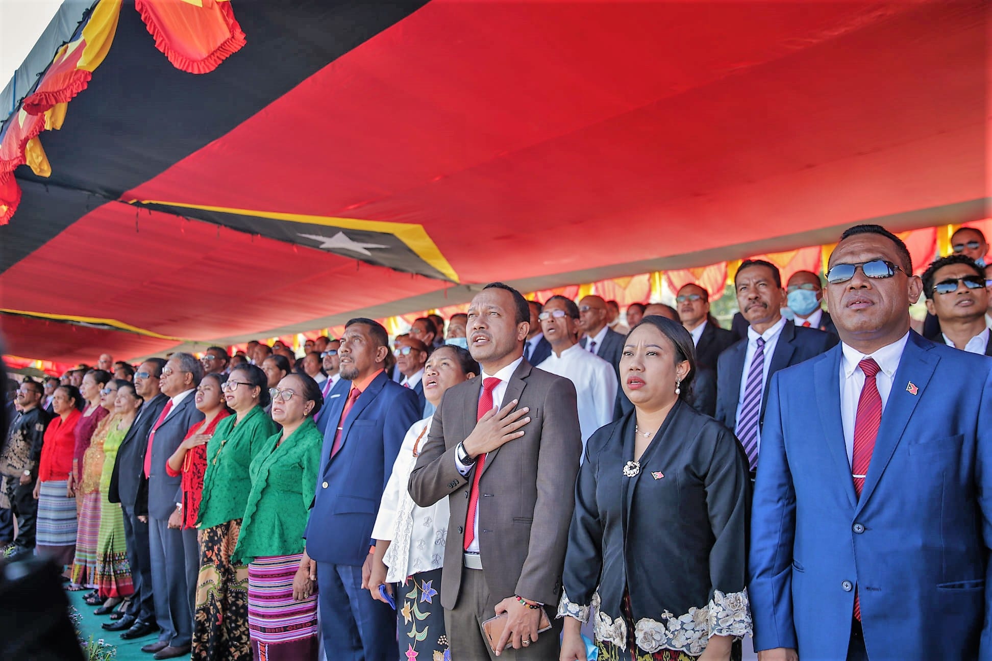 Rows of officials and guests in suits and traditional woven cloth standing under a red awning, hands on hearts, with a flag hanging overhead