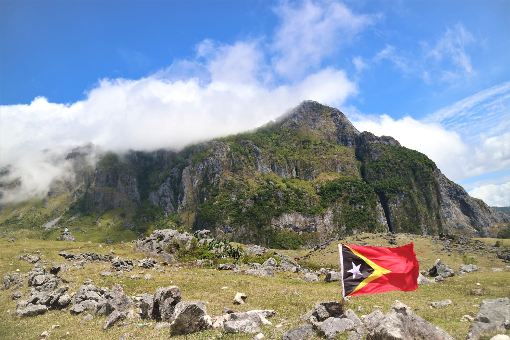 The red, yellow and black flag of Timor-Leste flying on a rocky grass slope below a steep green mountain wreathed in cloud