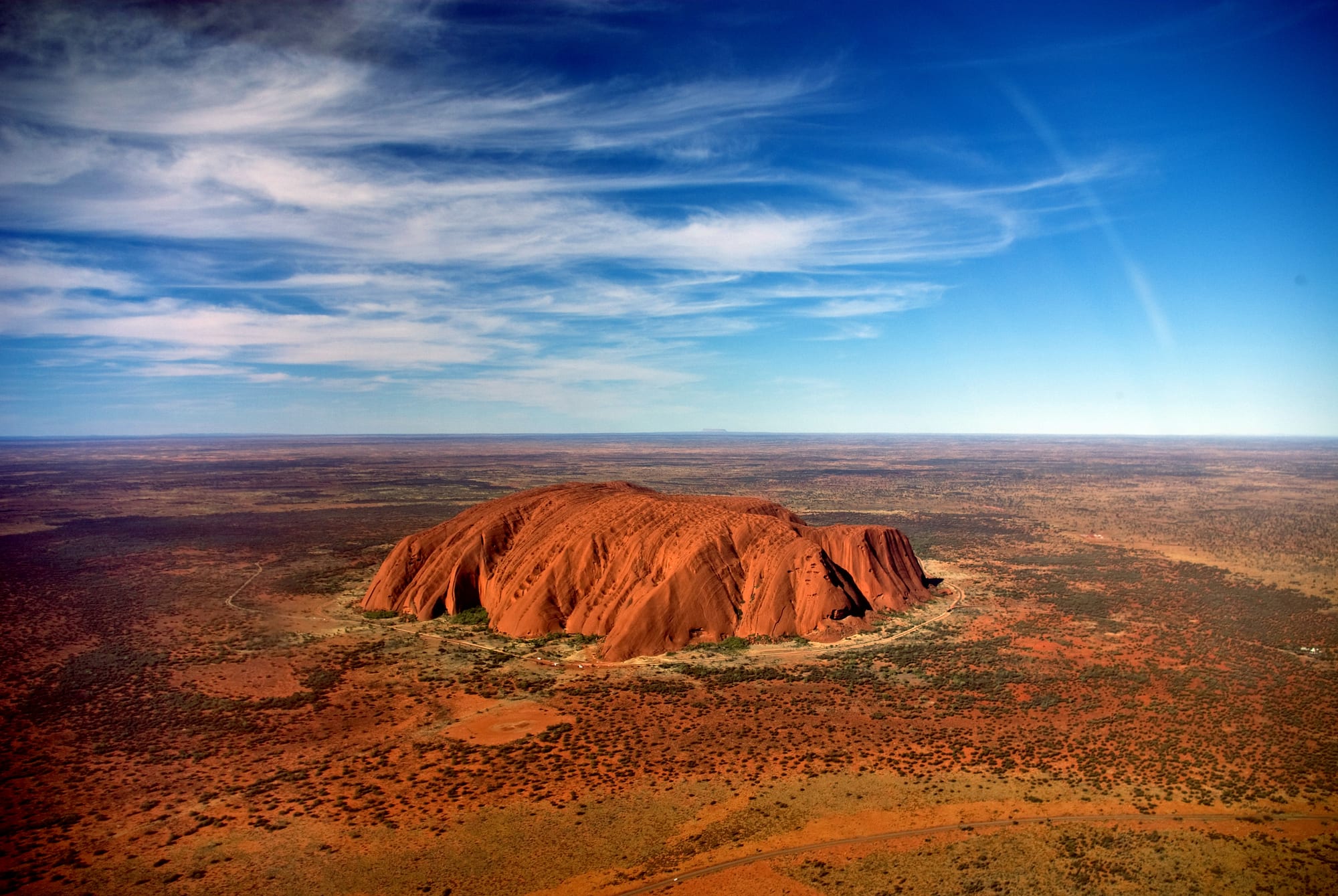 An aerial view of a huge red rock with deeply grooved sides rising alone from a flat red plain dotted with scrub, under a blue sky with streaky white cloud
