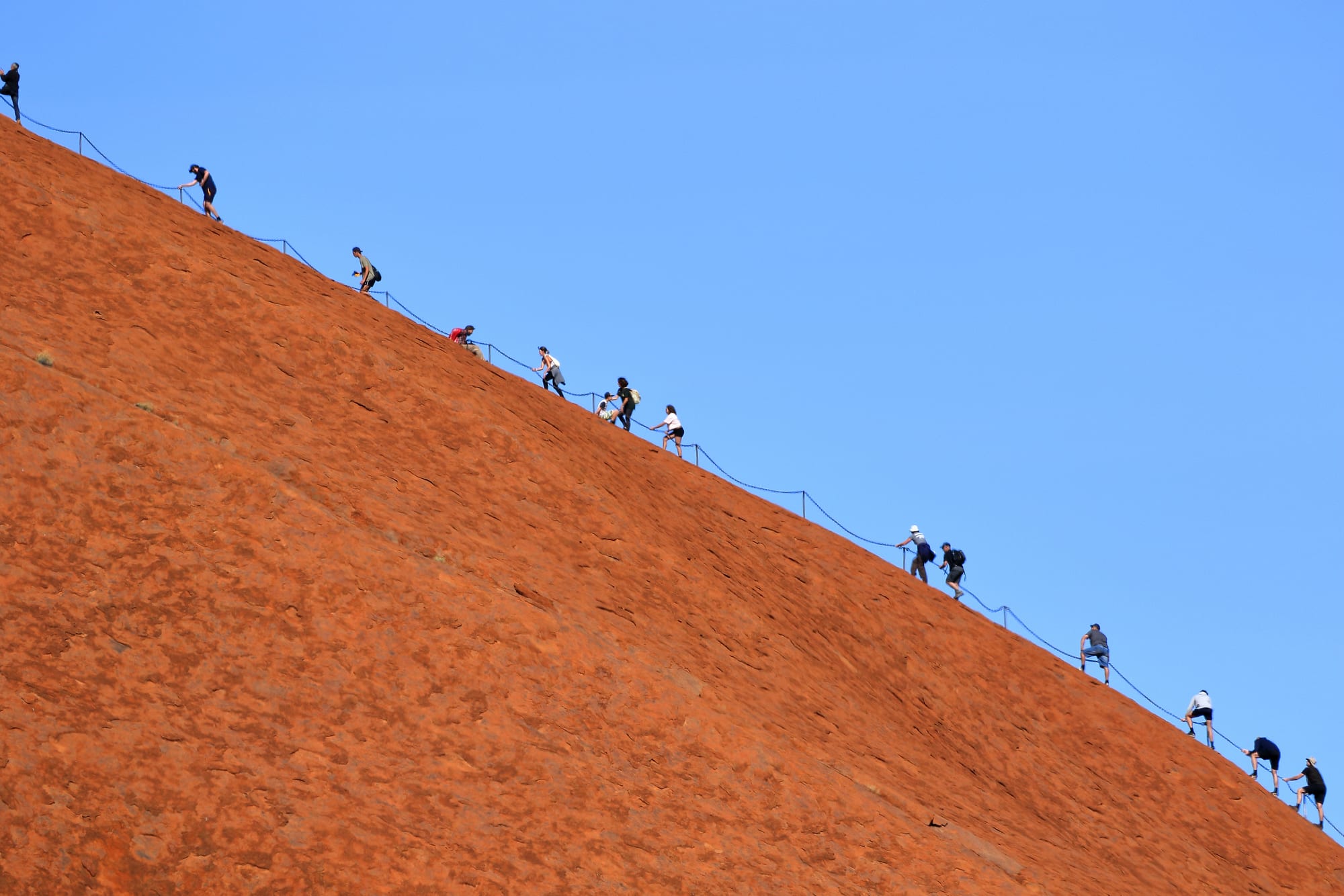 A line of small figures climbing a steep slope of red rock, holding a chain handrail against a clear blue sky