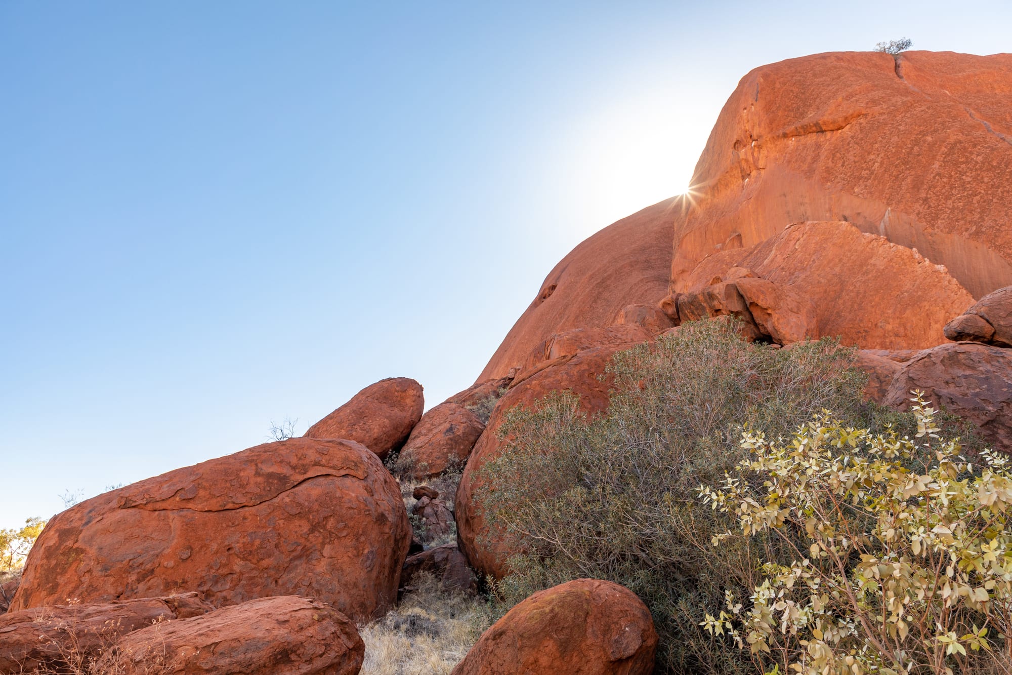 Close view of smooth, rounded orange-red rock faces and fallen boulders with grey-green shrubs, the sun peeking over the rock