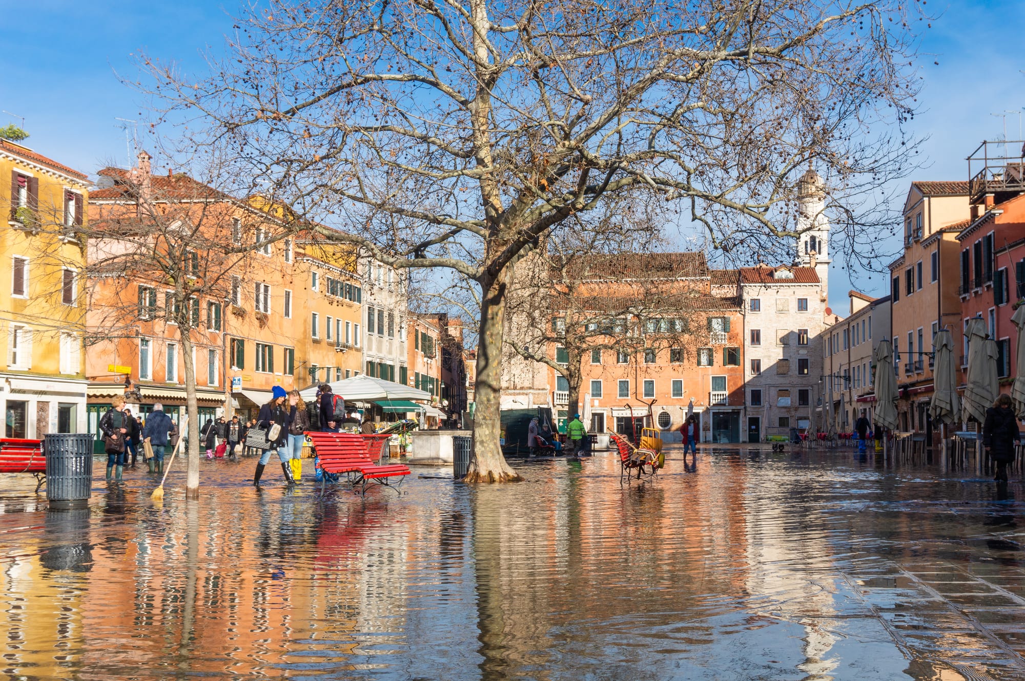 A city square covered in shallow water that reflects the orange and yellow houses around it, with people in rubber boots wading past a bare plane tree