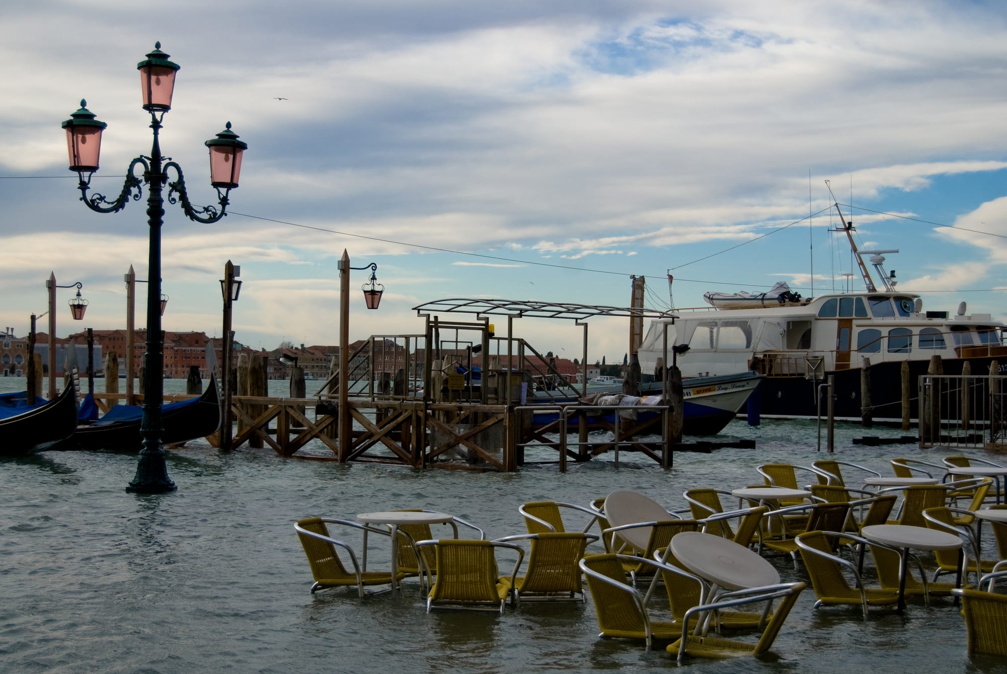 Café chairs and tables standing in shallow water beside a lagoon waterfront, with an ornate street lamp, gondolas and a moored motor yacht