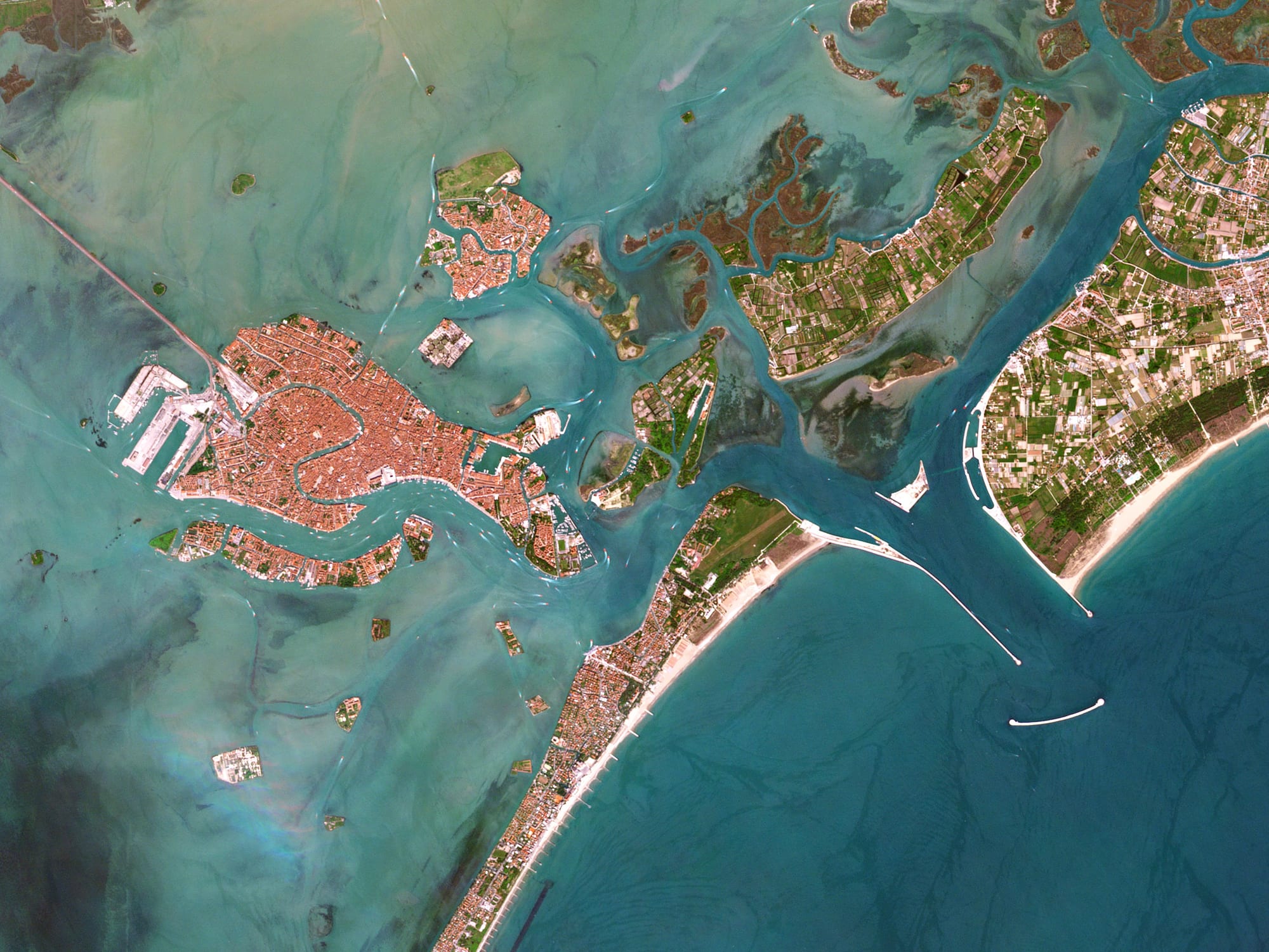 A satellite view of a red-roofed island city in a turquoise lagoon, with a long sandbar and breakwaters at an opening to the sea