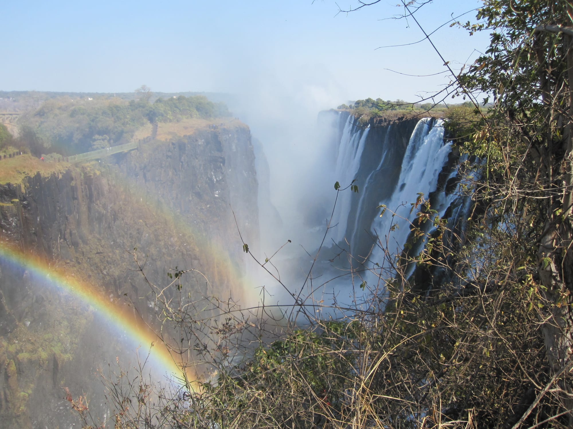 White water pouring over a dark cliff into a deep gorge filled with mist, with a rainbow arching across the spray in the foreground