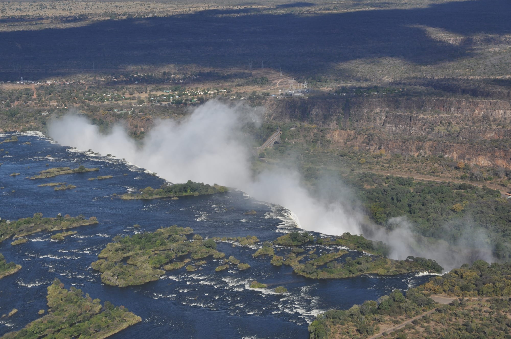 An aerial view of a wide river dotted with wooded islands, plunging into a deep crack in the land and sending up a long cloud of white spray