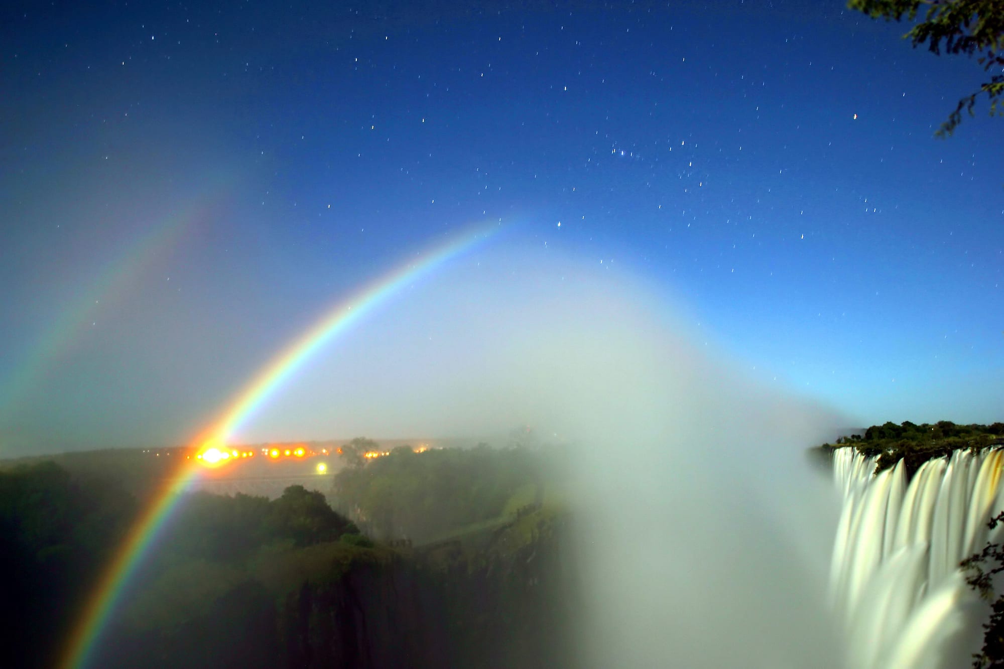 A pale double rainbow arching over a misty gorge under a starry night sky, with a floodlit waterfall on the right