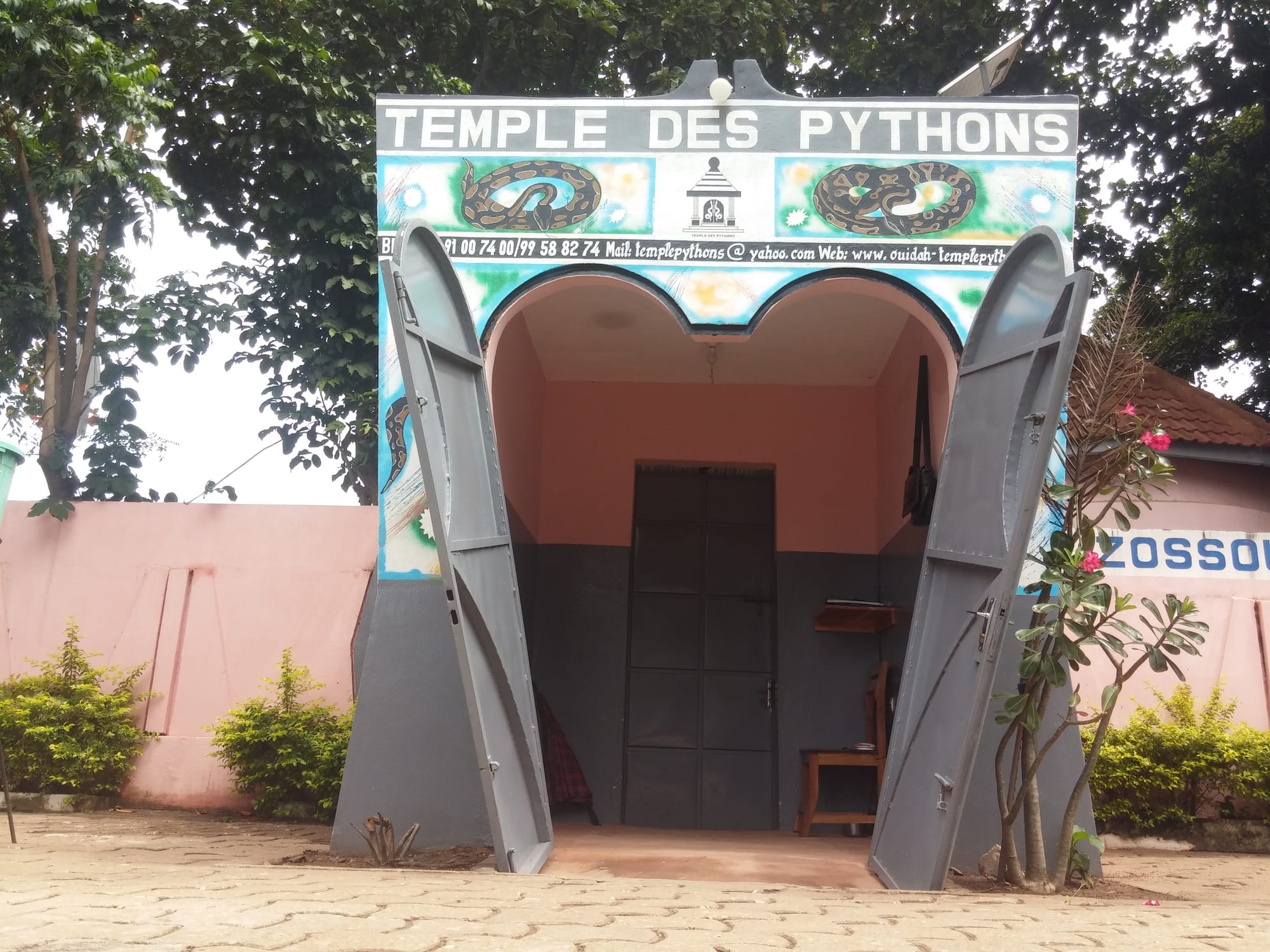 A small gatehouse painted with pictures of coiled pythons under the words Temple des Pythons, its grey metal doors open, set in a pink wall among trees