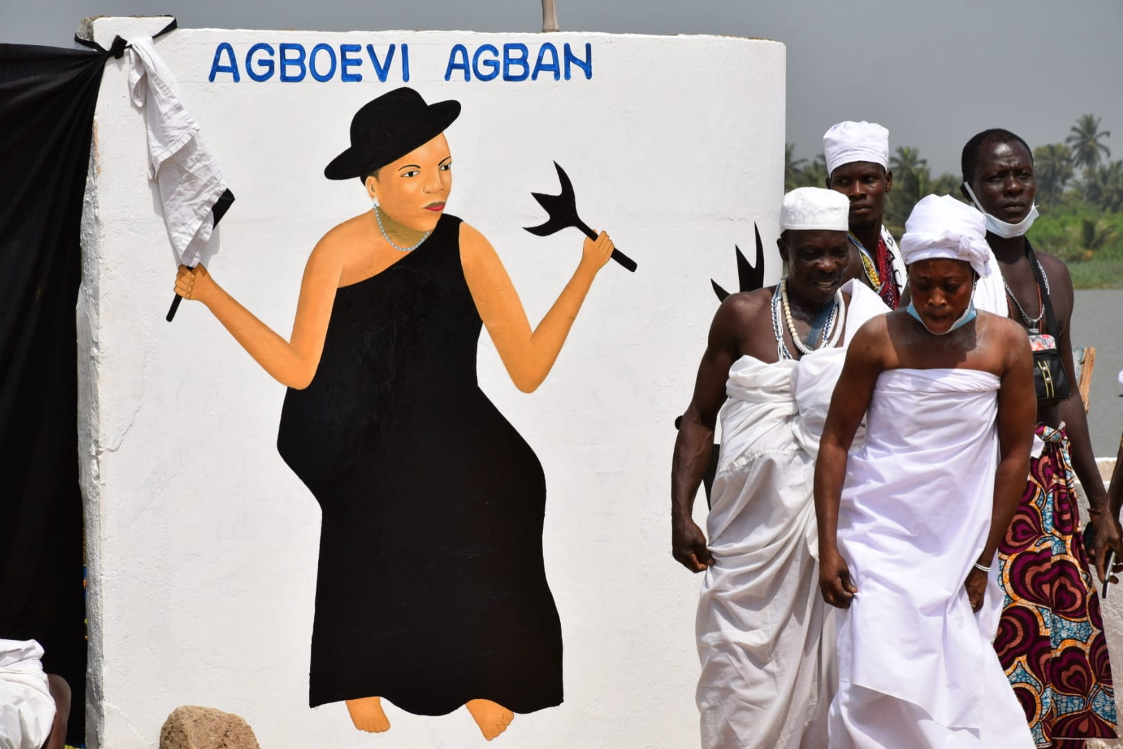 A man and a woman wrapped in white cloth and wearing white caps walk past a wall painted with a woman in a black dress holding a white cloth and a dark axe-shaped object, beside a lagoon