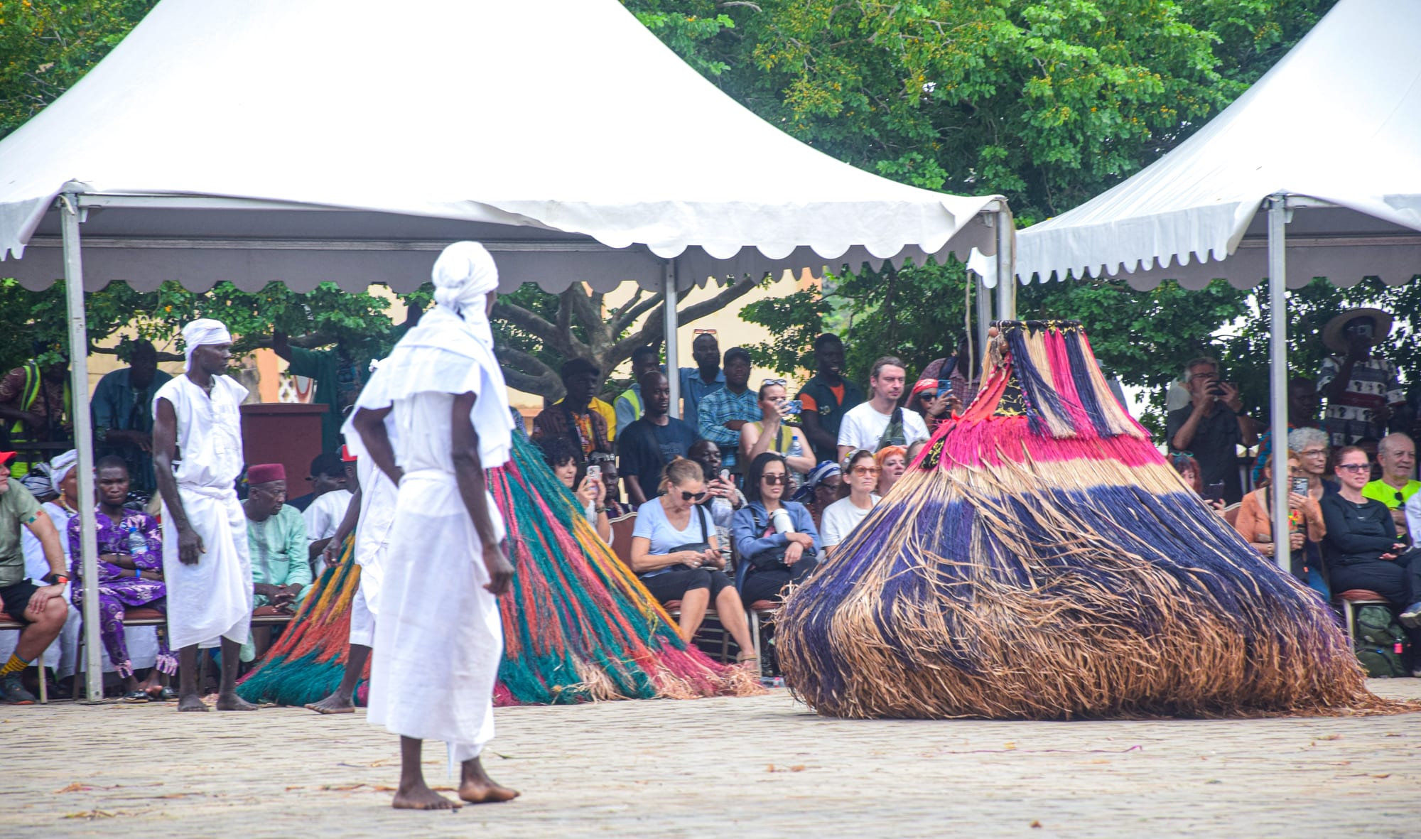 Two tall, cone-shaped figures covered in long strands of dyed raffia in pink, blue and green, performing on a paved square watched by people in white robes and a seated crowd under tents