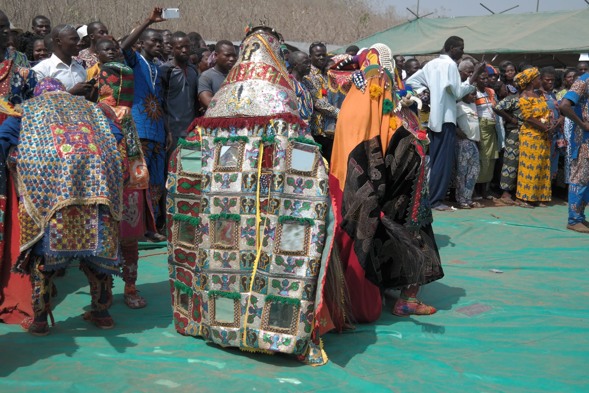 Several masked dancers in heavy, richly embroidered costumes decorated with small mirrors, surrounded by a dense crowd of onlookers