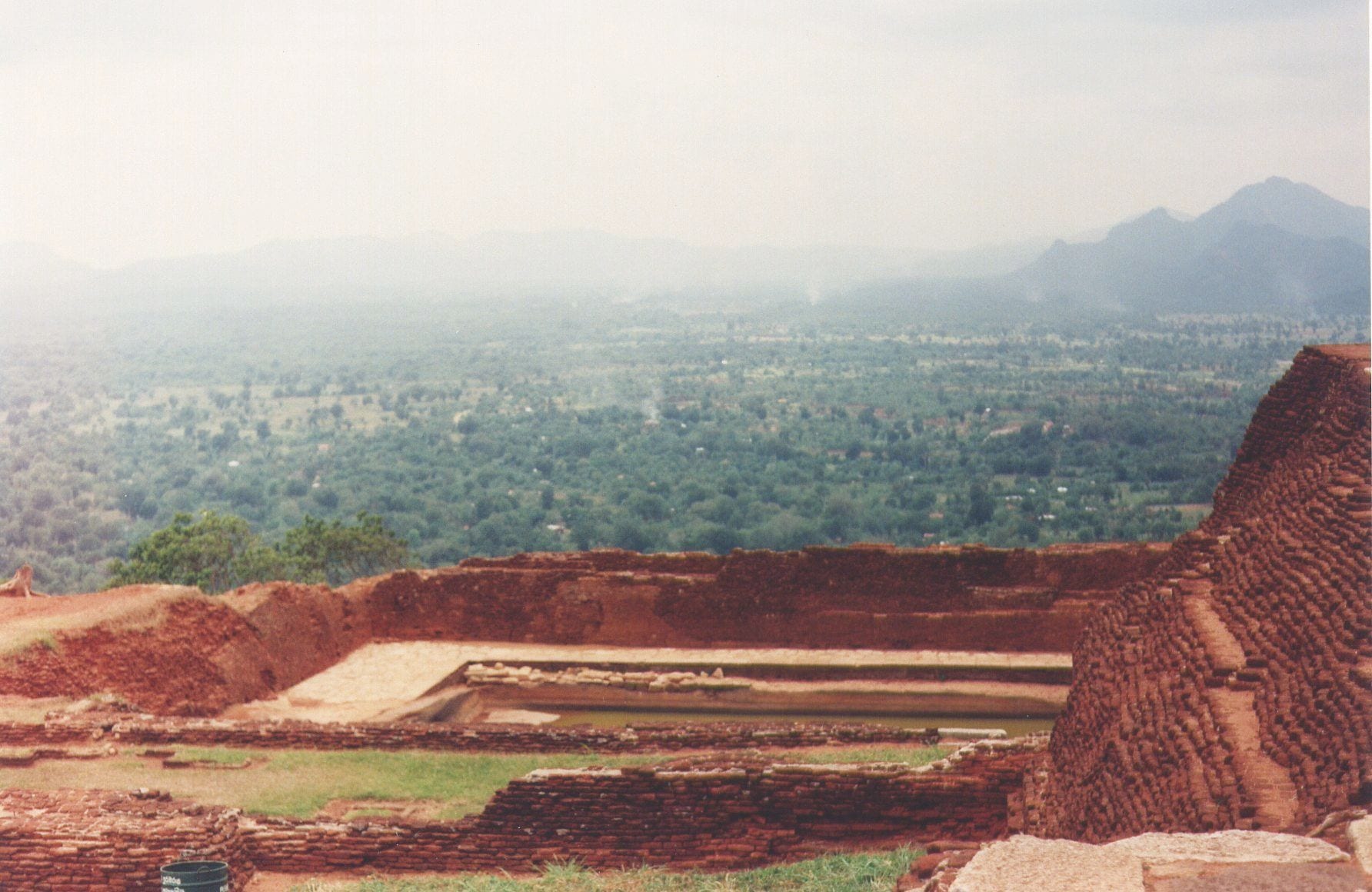 Red brick foundations and a rectangular rock-cut pool on a summit, looking out over a hazy forested plain to distant hills