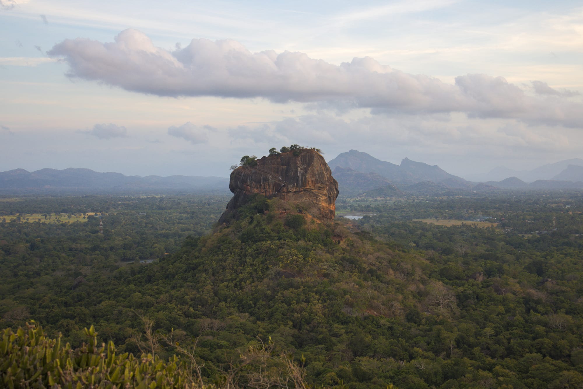 A sheer-sided, flat-topped rock rising out of a forested hill above a wide green plain, with mountains on the horizon