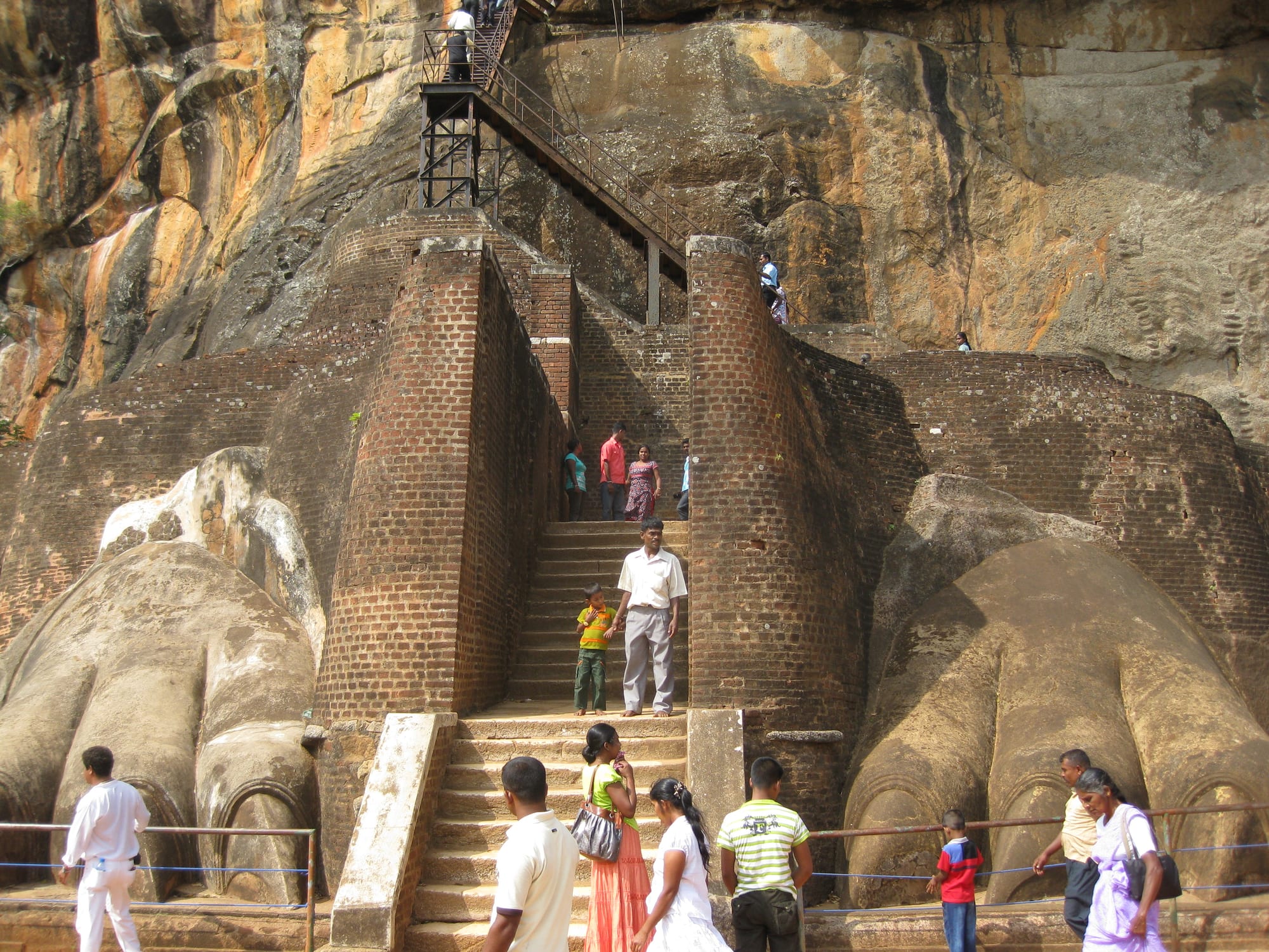 A brick staircase climbing between two enormous carved stone lion paws at the foot of a sheer rock face, with visitors on the steps