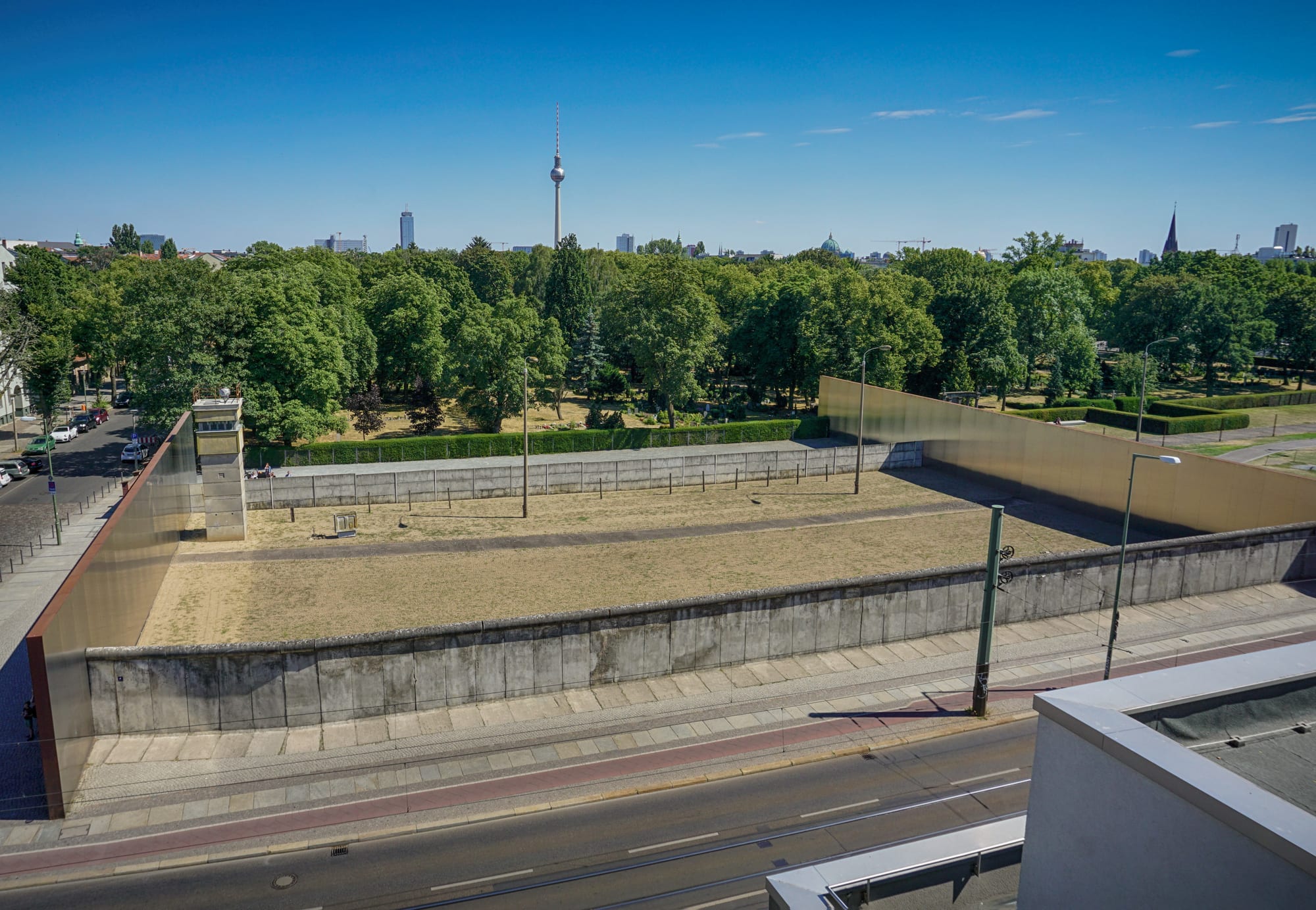 A preserved stretch of grey concrete wall beside a street, with an empty sandy strip, a second wall and a watchtower behind it, and a tall television tower on the skyline