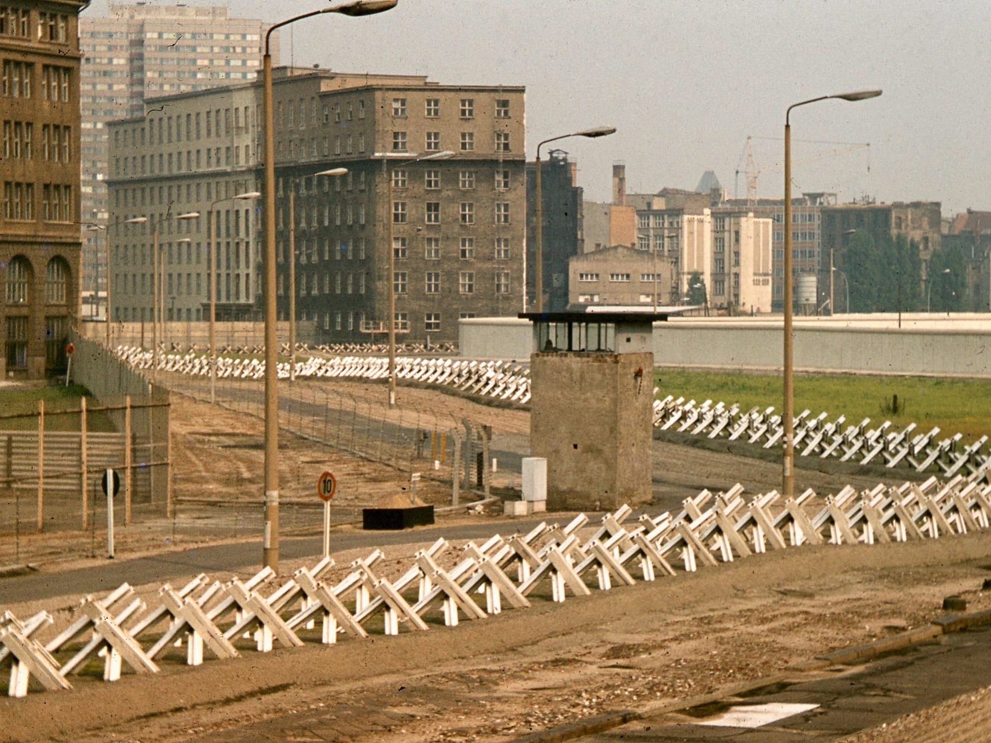 A wide empty strip of sand lined with rows of white crossed-steel anti-tank obstacles, a concrete guard tower and a long white wall, with apartment blocks behind