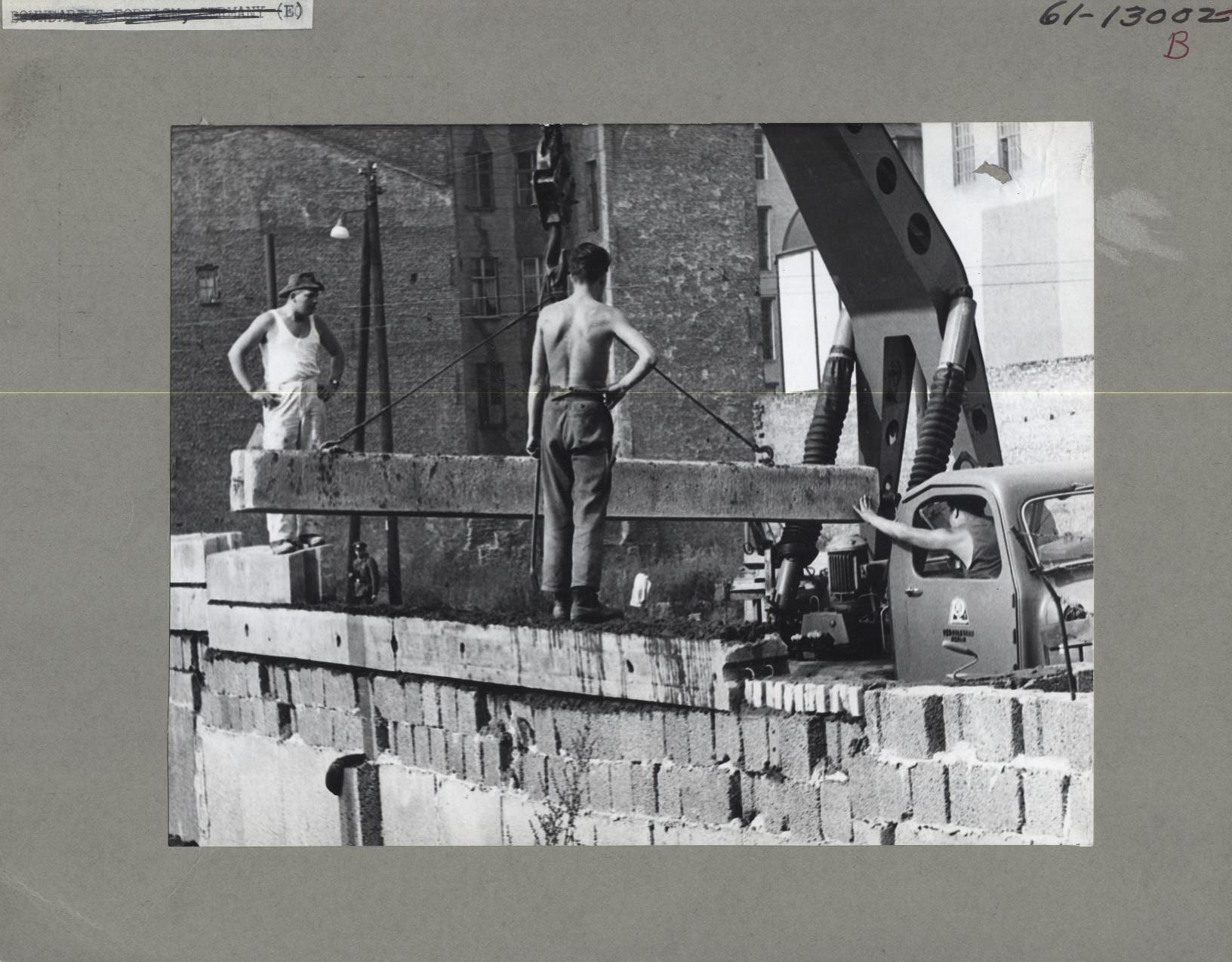 A black and white photograph of workers in vests standing on a wall of concrete blocks as a crane lowers a long concrete beam onto it