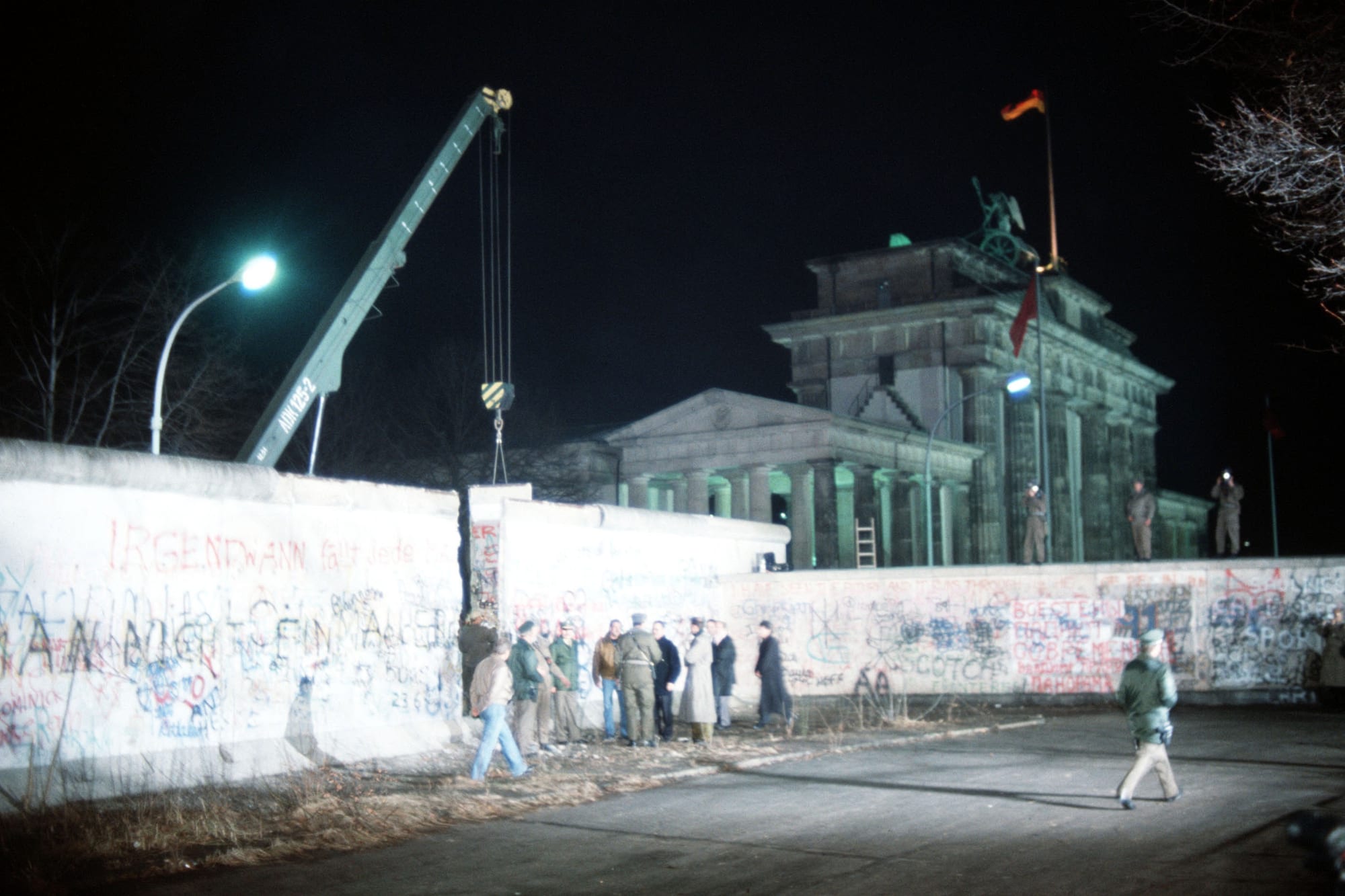At night, a crane stands beside a graffiti-covered wall with a gap in it, with a crowd gathered in front and the floodlit Brandenburg Gate behind
