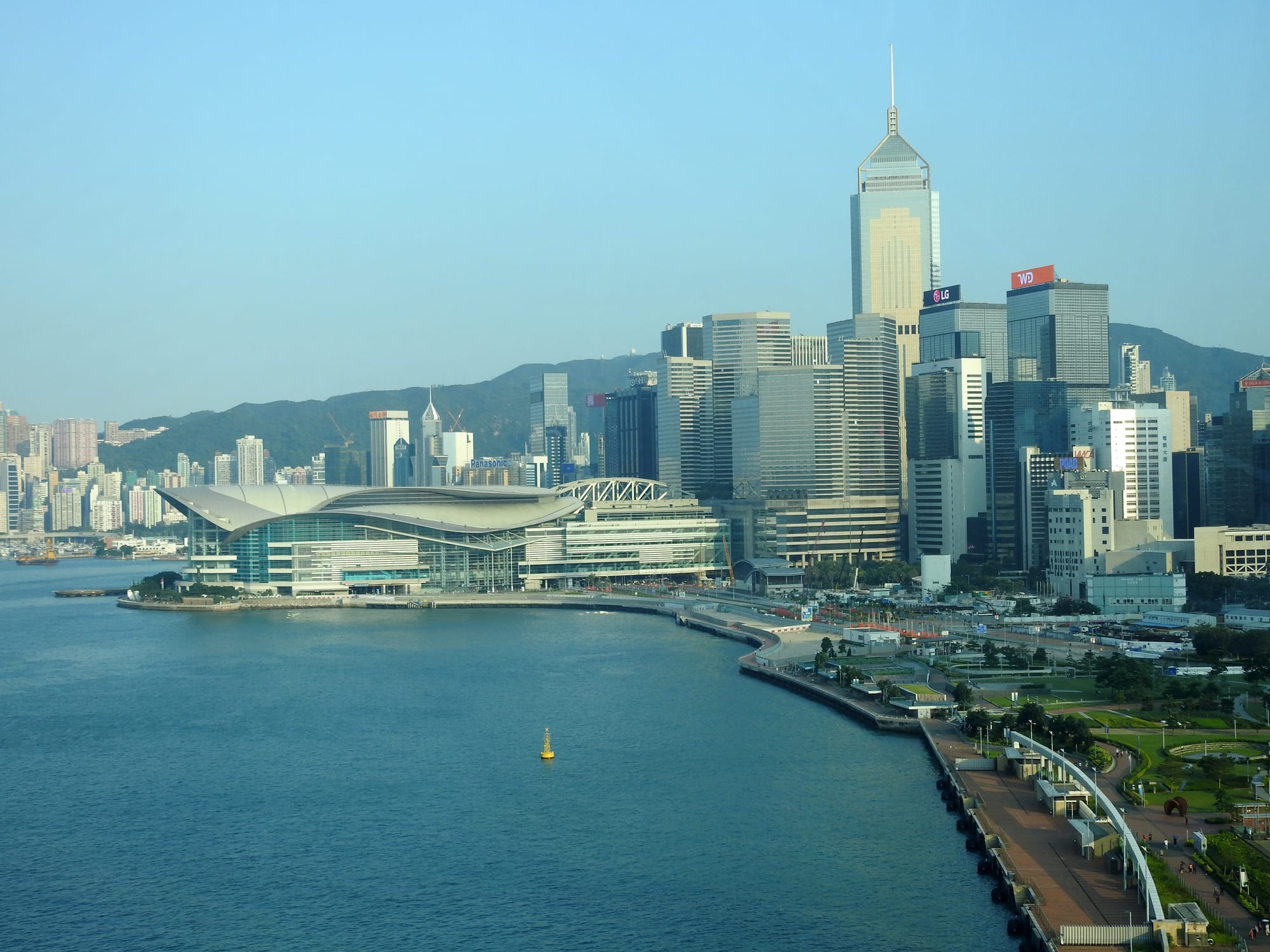 A curved glass exhibition hall on a promontory in a blue harbour, with office towers and green hills behind