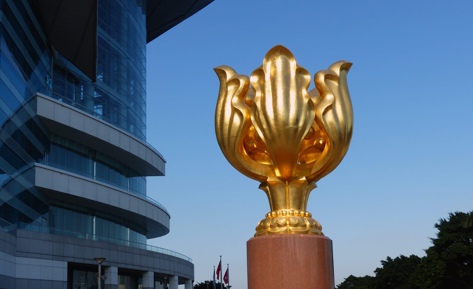 A giant gilded flower sculpture on a red granite pillar beside a curved glass building