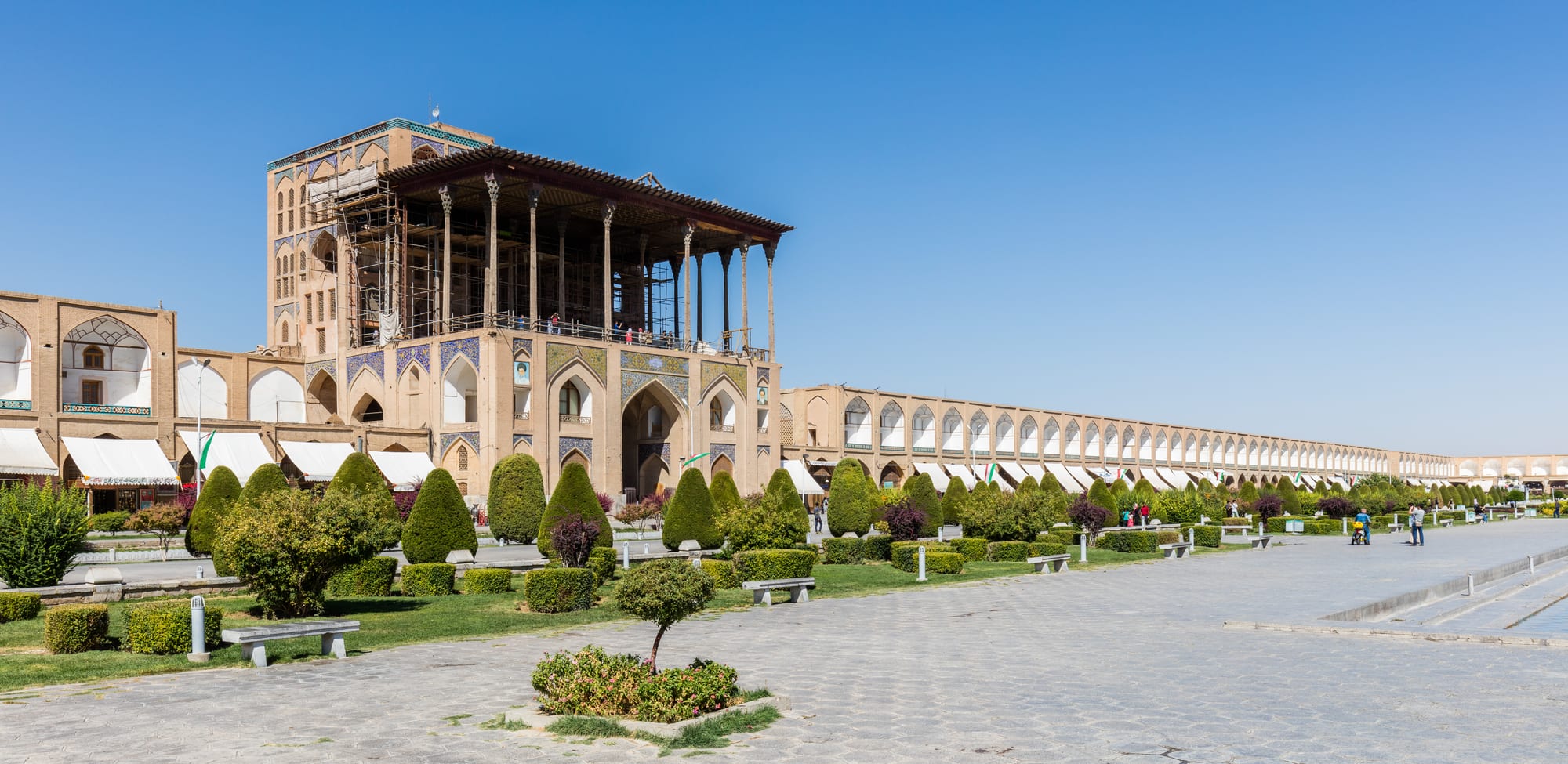 A tall palace with a high wooden-columned terrace, set into a long two-storey arcade along the edge of a paved square with clipped trees