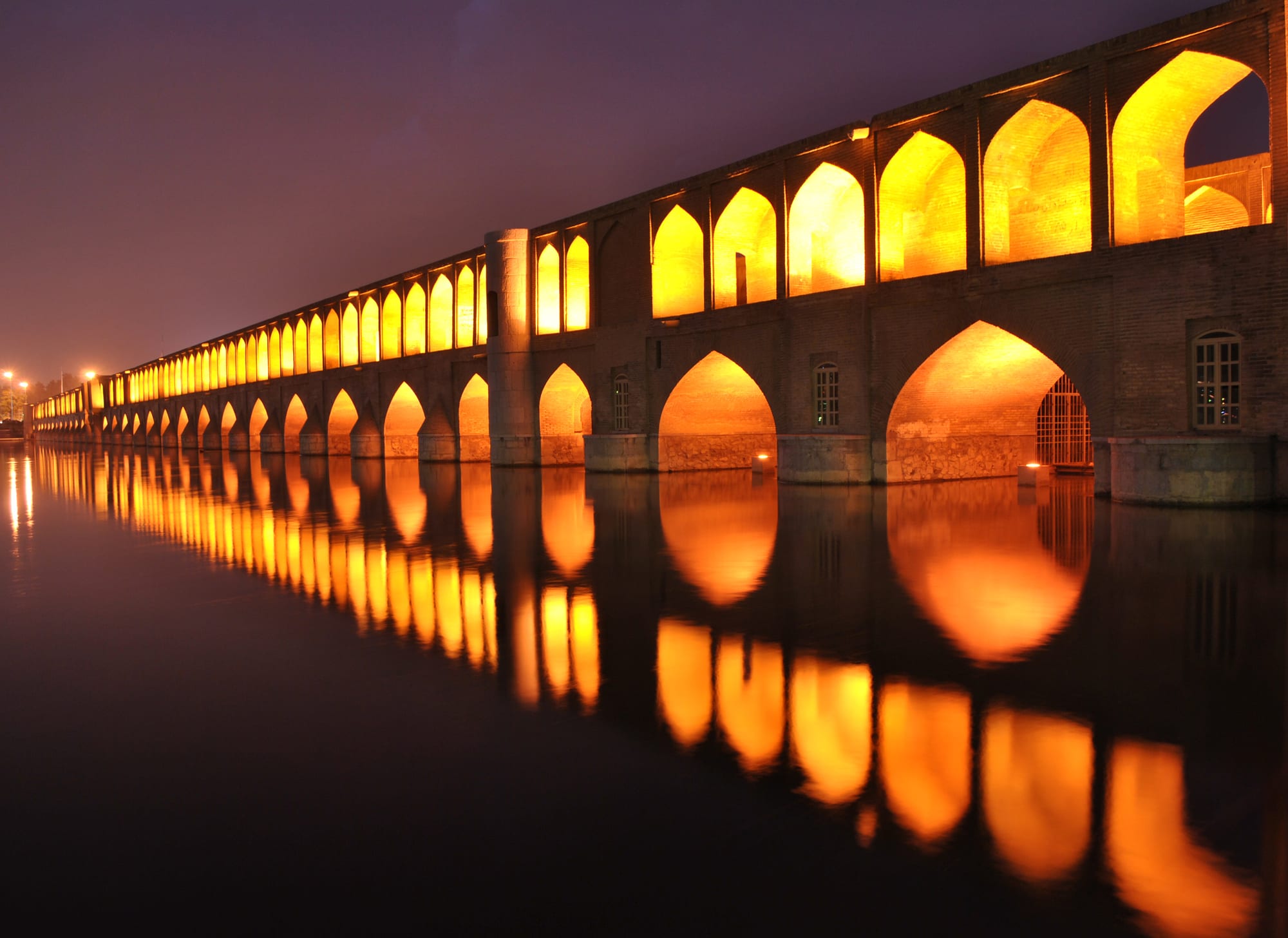 A long two-tier arched bridge lit gold at night, its arches reflected in still water