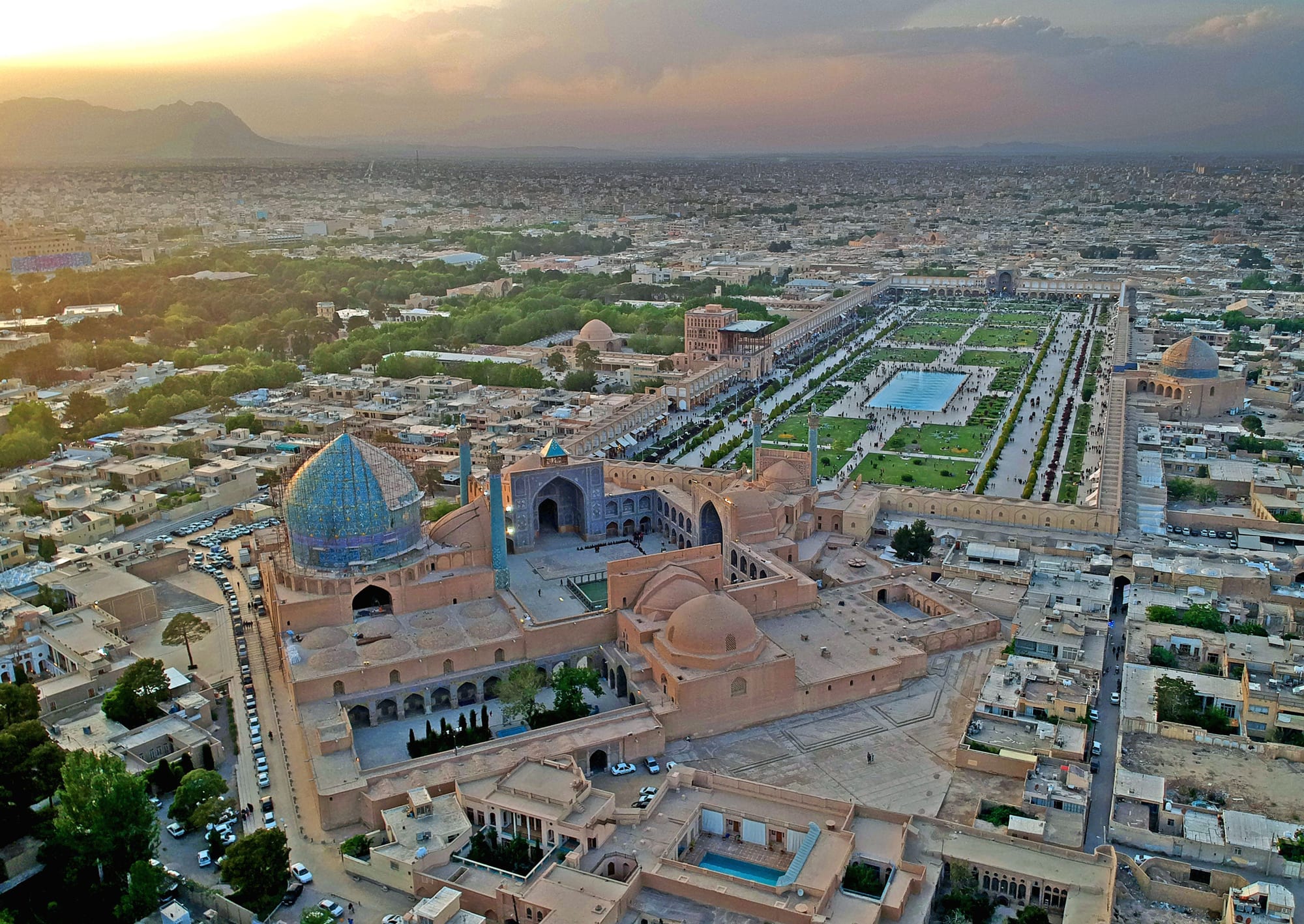An aerial view of a vast rectangular square with lawns and a long pool, a large blue-tiled mosque dome in the foreground and a second dome on the far side