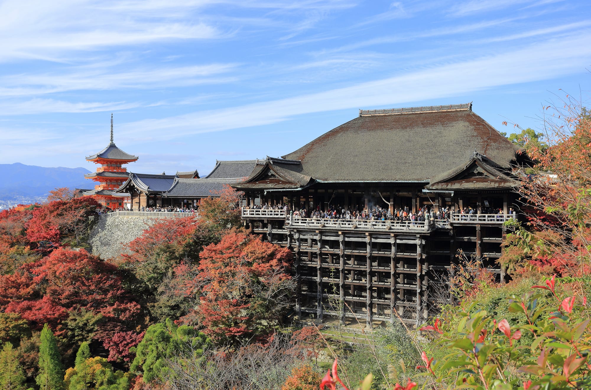 A large wooden temple hall on a tall wooden scaffold above autumn trees in red and orange, with a red pagoda to the left and mountains behind