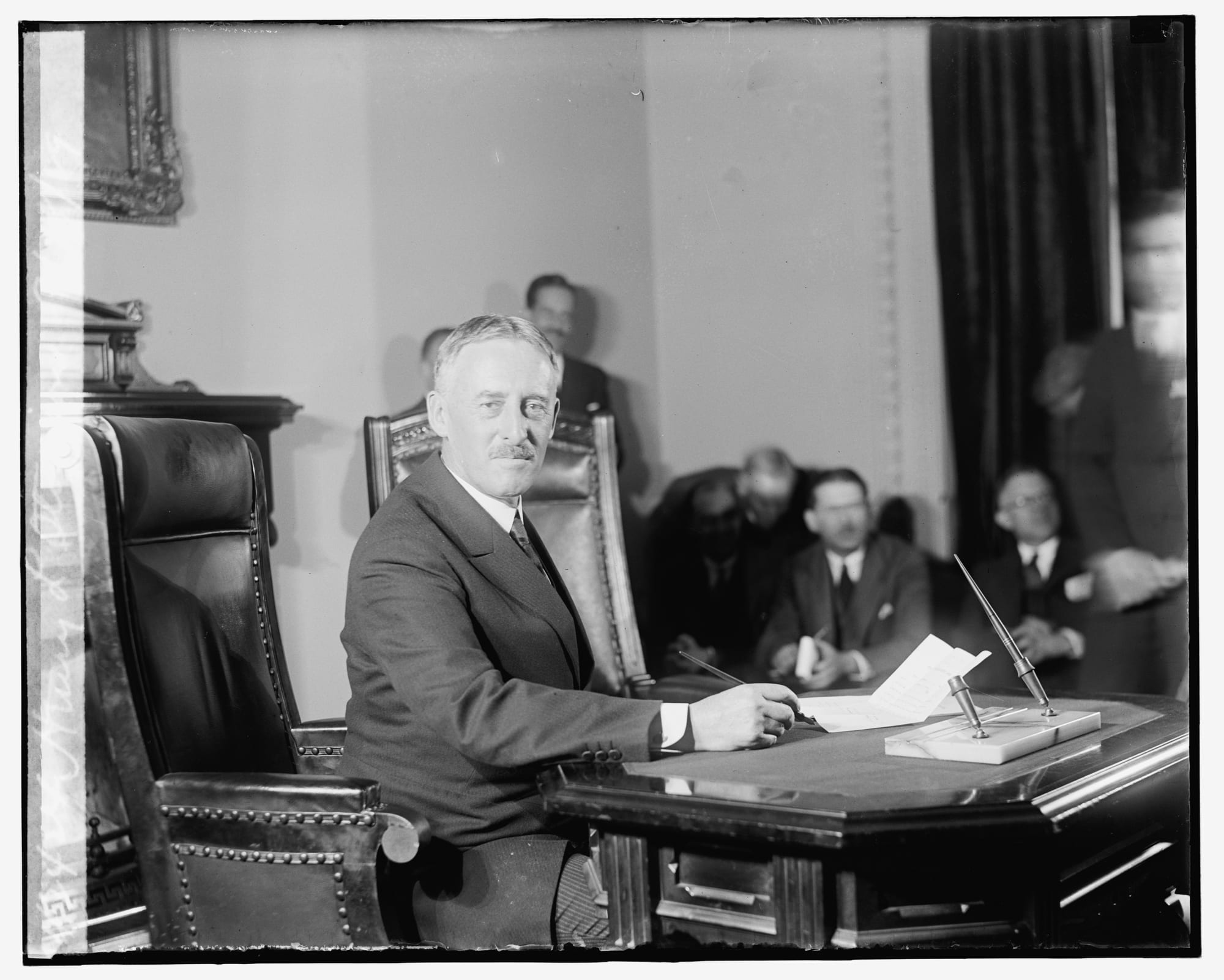 A black-and-white photograph of a grey-haired man with a moustache seated at a large desk, with other men in suits behind him