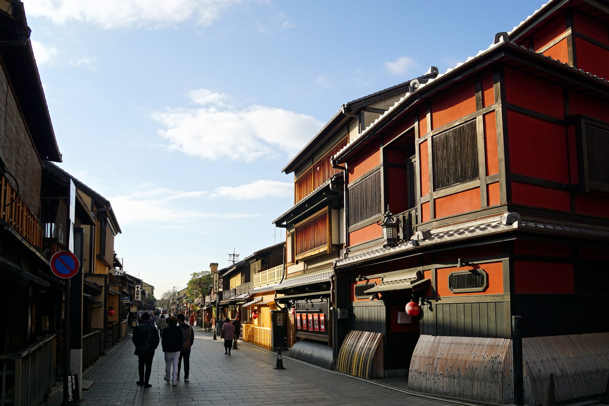 A paved street lined with two-storey wooden townhouses with red walls, slatted windows and hanging lanterns, with people walking under a blue sky