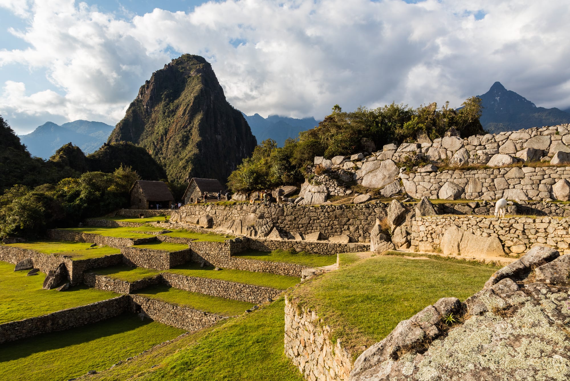 Grassy stone terraces and walls of fitted grey stone below a steep, pointed green mountain under a cloudy sky