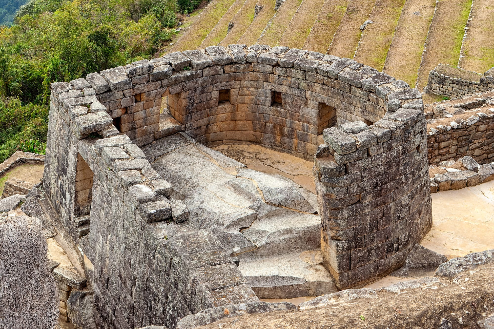 A curved stone tower of finely cut, tightly fitted blocks with trapezoidal windows, enclosing a carved rock