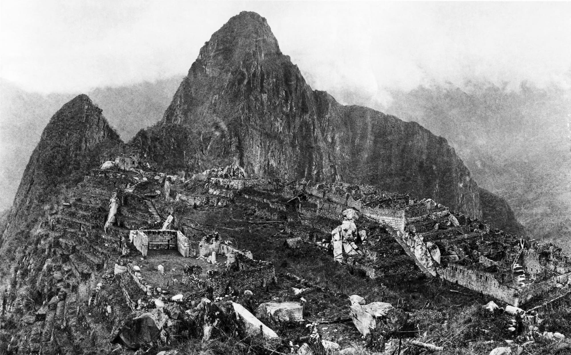 A black and white photograph of overgrown stone ruins and terraces on a ridge below a steep, pointed peak