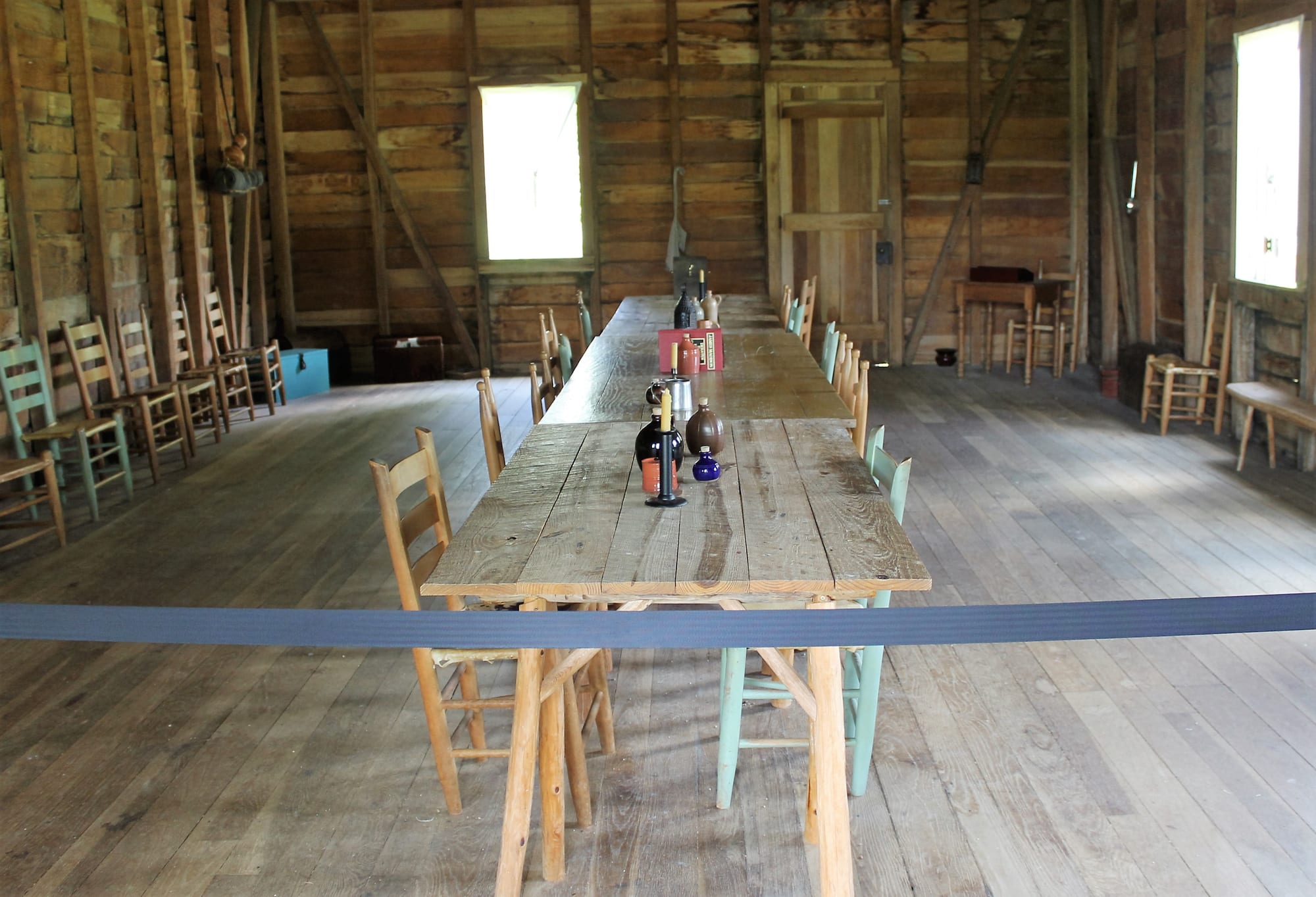A long plain wooden table with candles and bottles, surrounded by simple chairs, in a bare plank-walled room